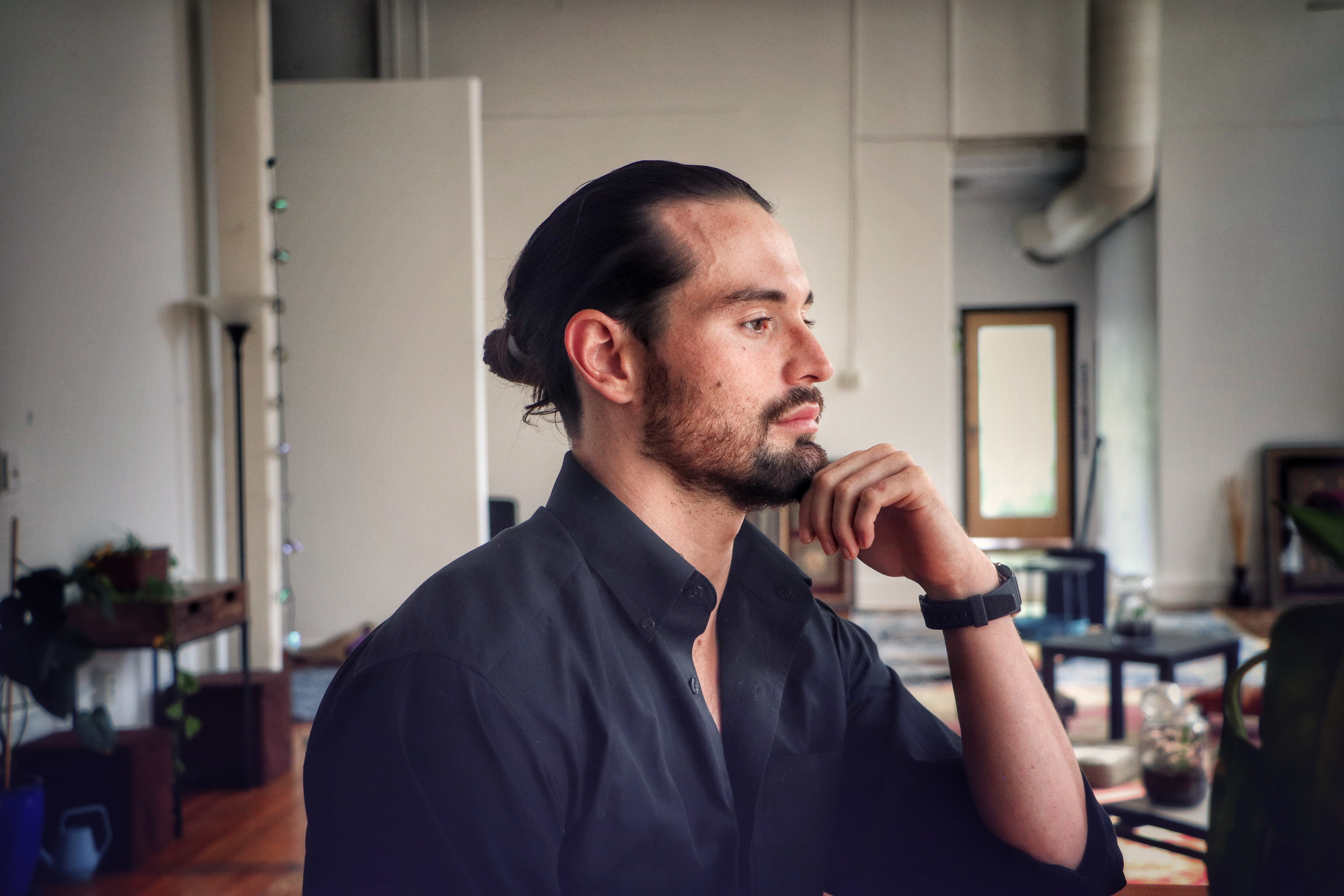 Side profile of a man with dark hair tied back, wearing a dark shirt, sitting in an office, resting his chin on his hand.