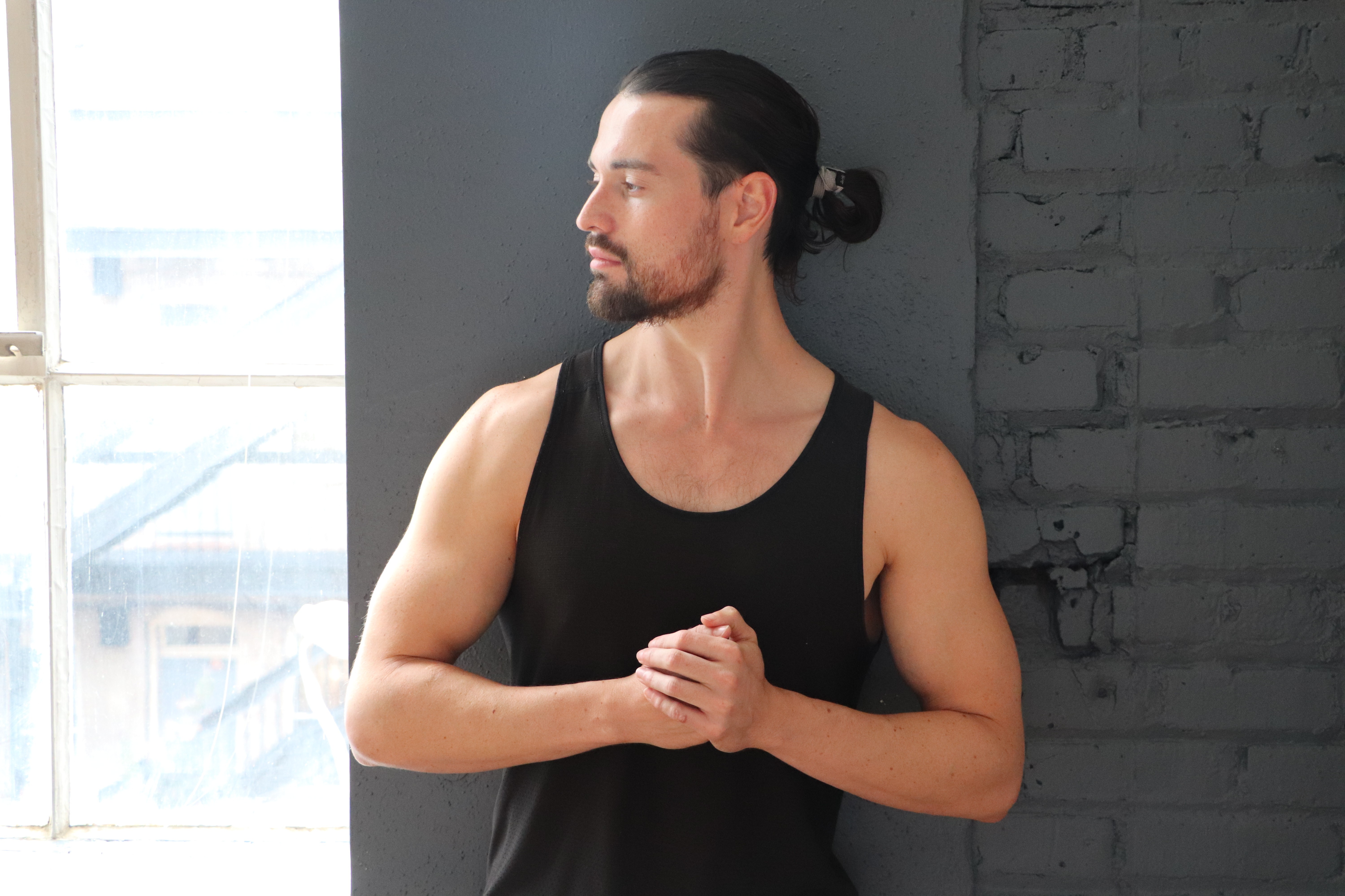 Man with long dark hair tied back, wearing a black sleeveless shirt, standing indoors near a window and gray brick wall.