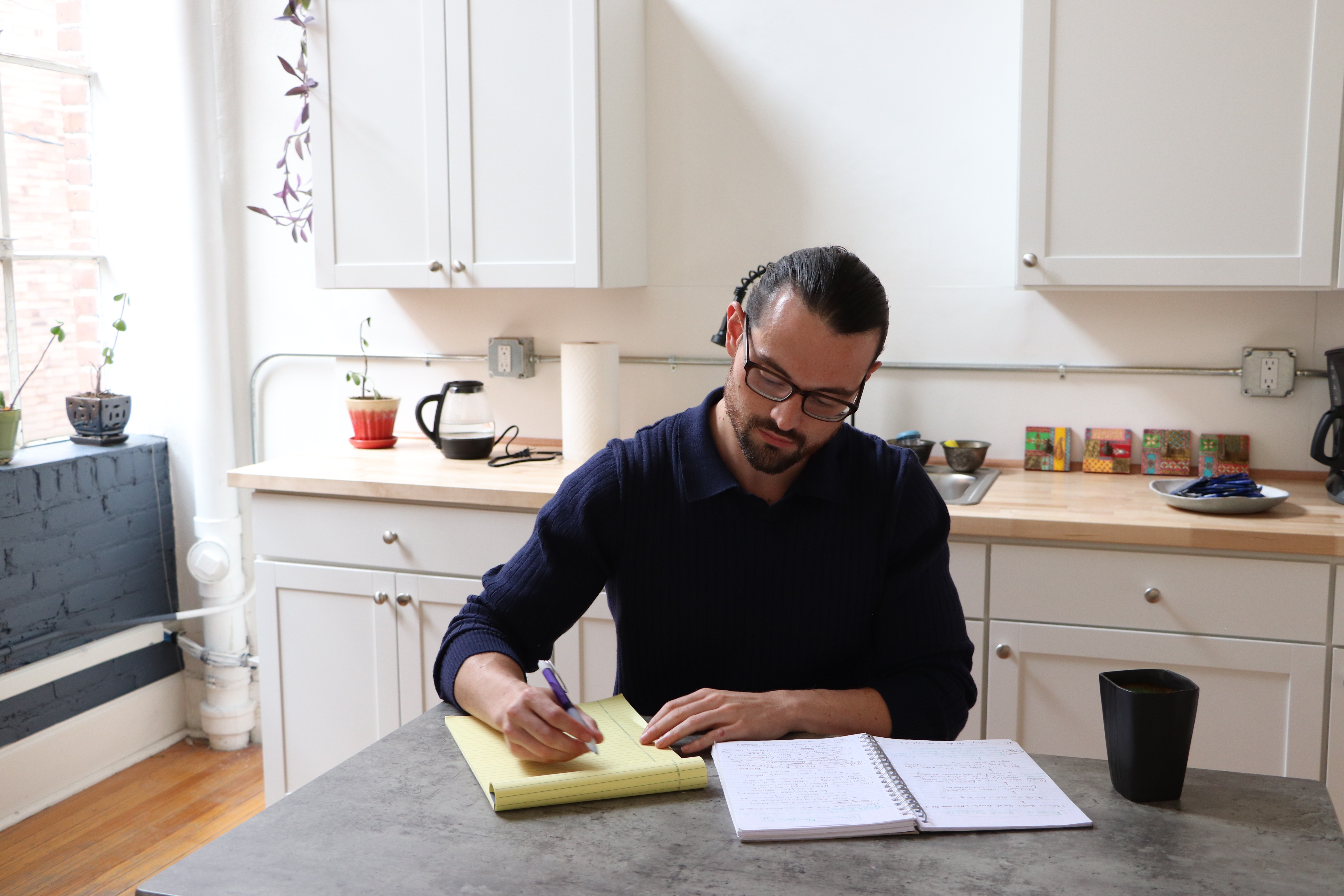 Man with glasses writing on a yellow notepad at a kitchen counter, with open book and black cup nearby.