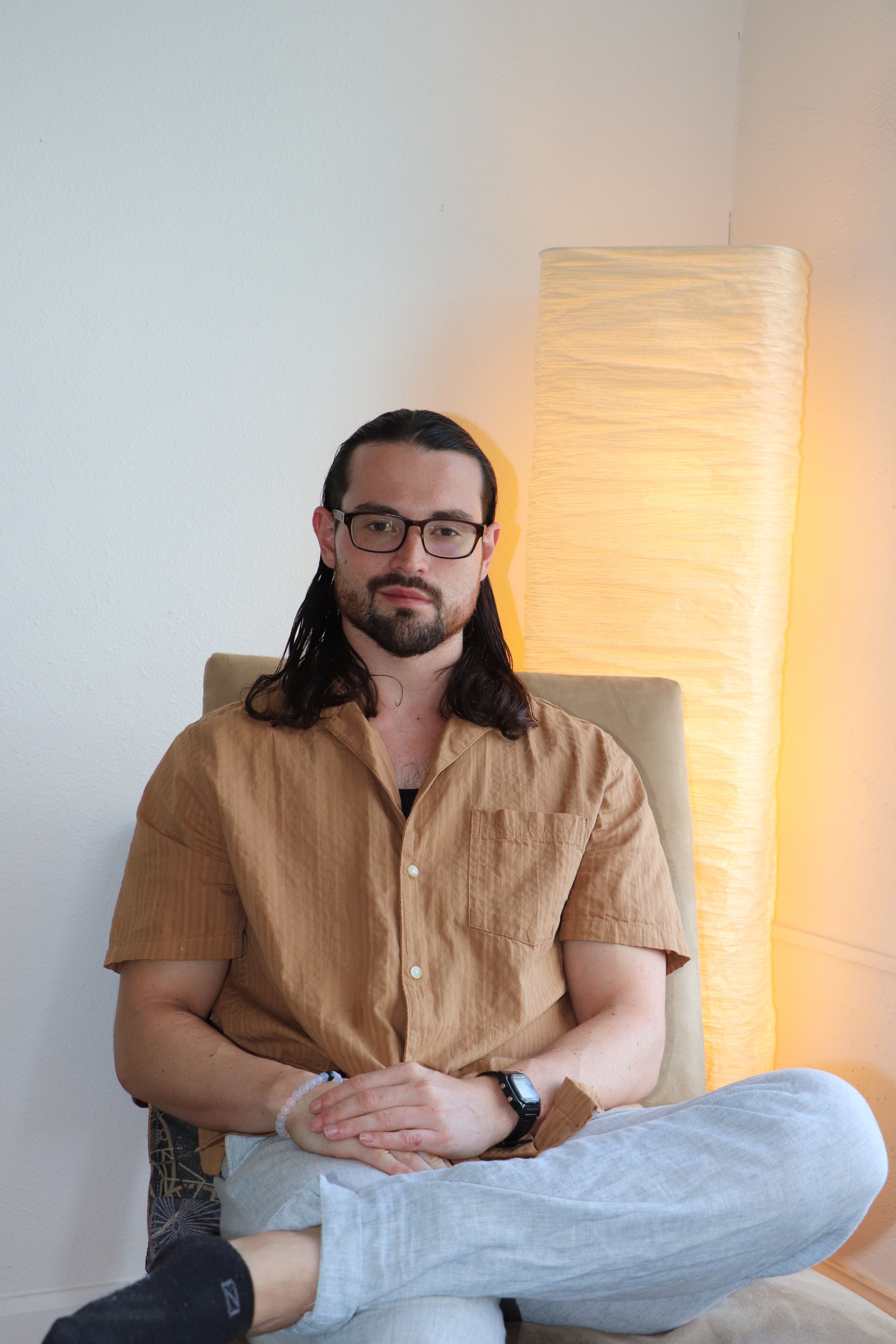 Woman with glasses and dark hair, wearing a brown shirt, sitting against a light-colored wall, looking at the camera.