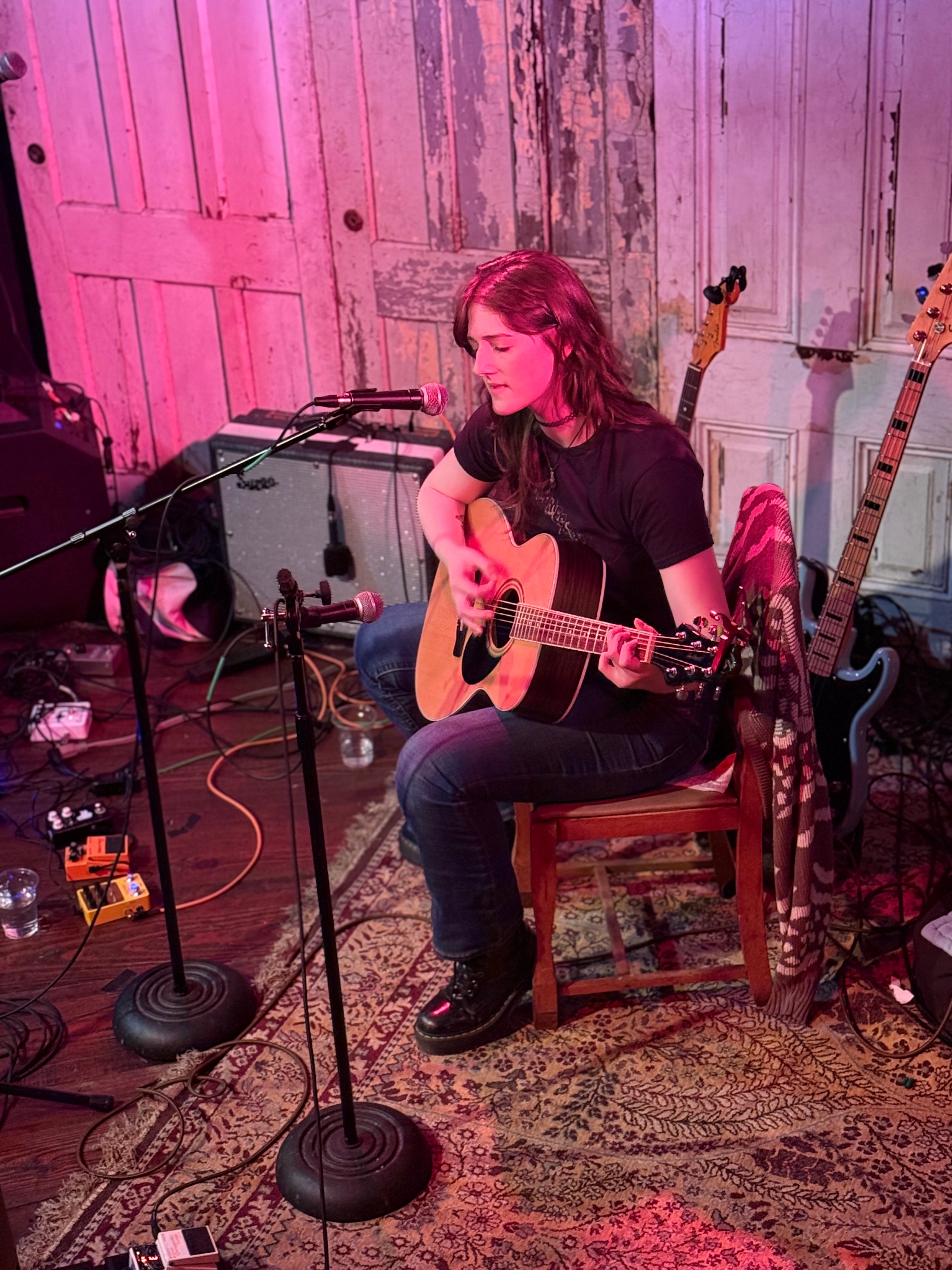 Young woman playing acoustic guitar on stage with microphones and musical equipment, pink lighting, and guitars in background.