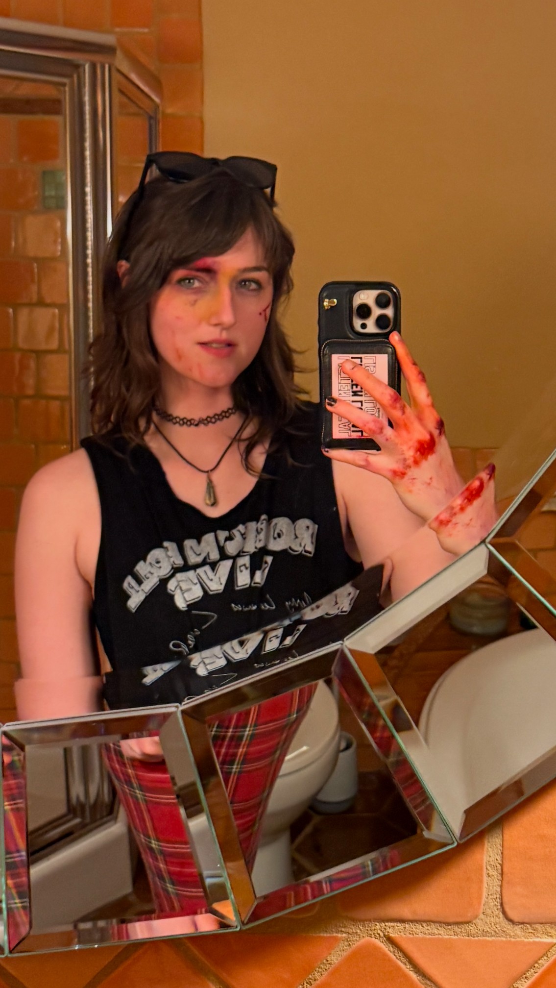 Young woman with dark hair and glasses on her head taking a mirror selfie, wearing a black tank top and plaid pants, with a brick wall background.