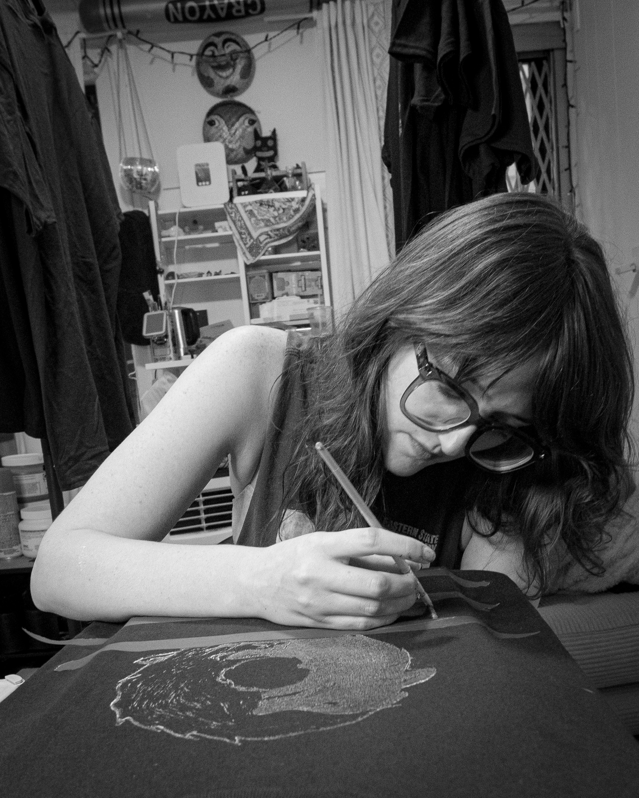 Person with glasses drawing on a table, cluttered room in background, black and white photo.