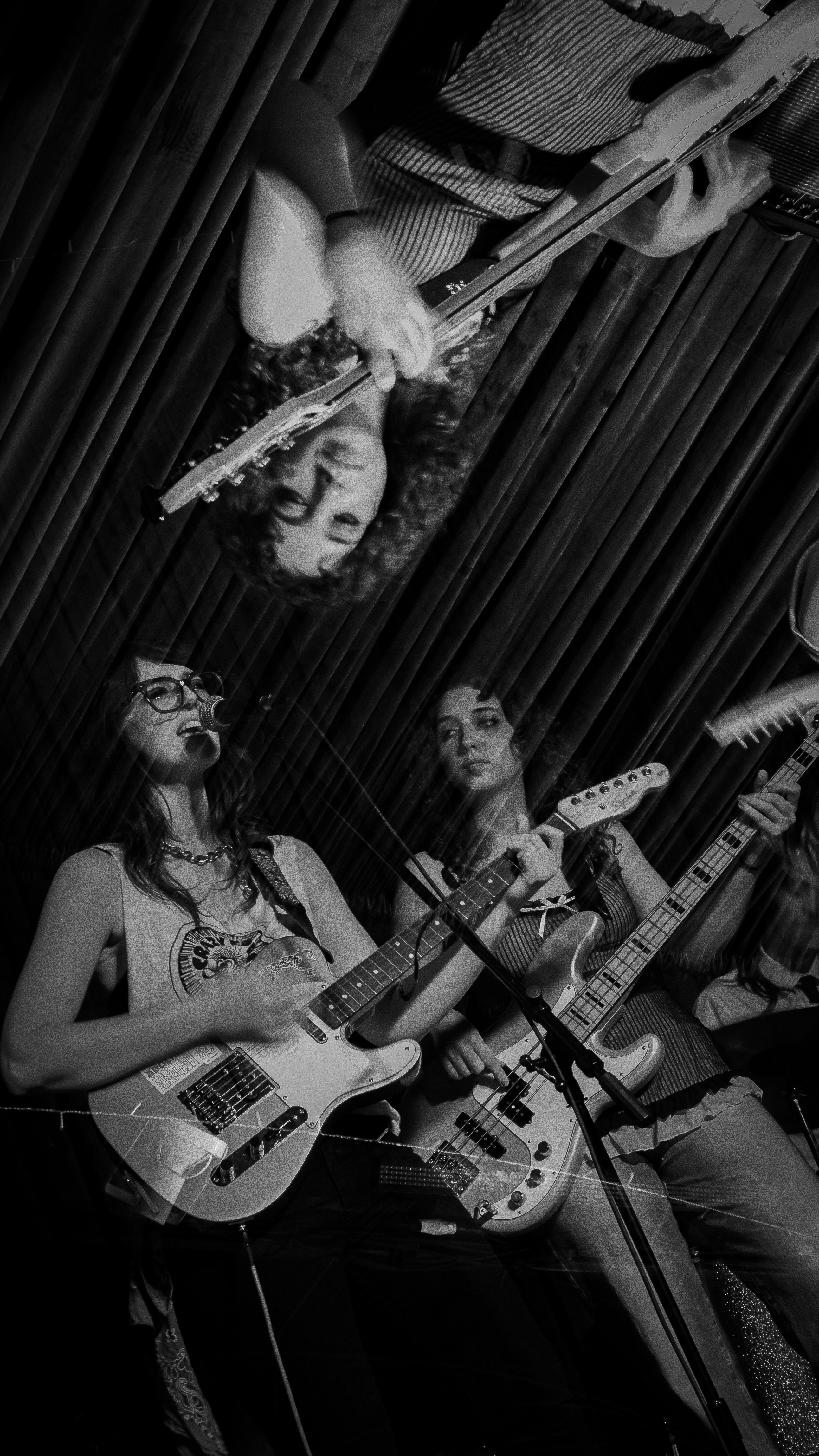 Two women playing guitars on stage with a curtain background, one singing and the other looking at her, in black and white.