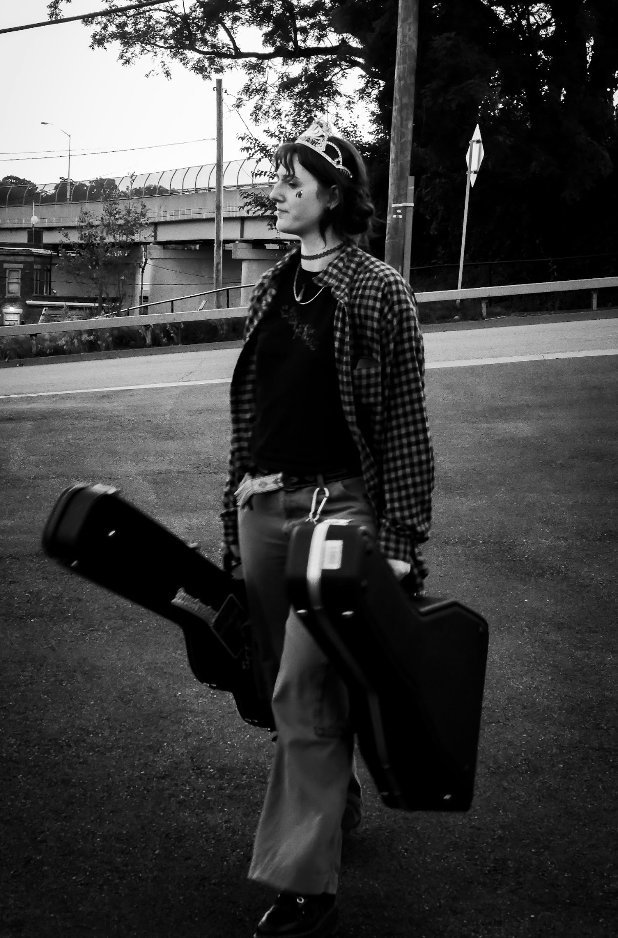 Person standing outdoors holding two guitar cases, wearing glasses and a checkered jacket, with trees and a building in background.