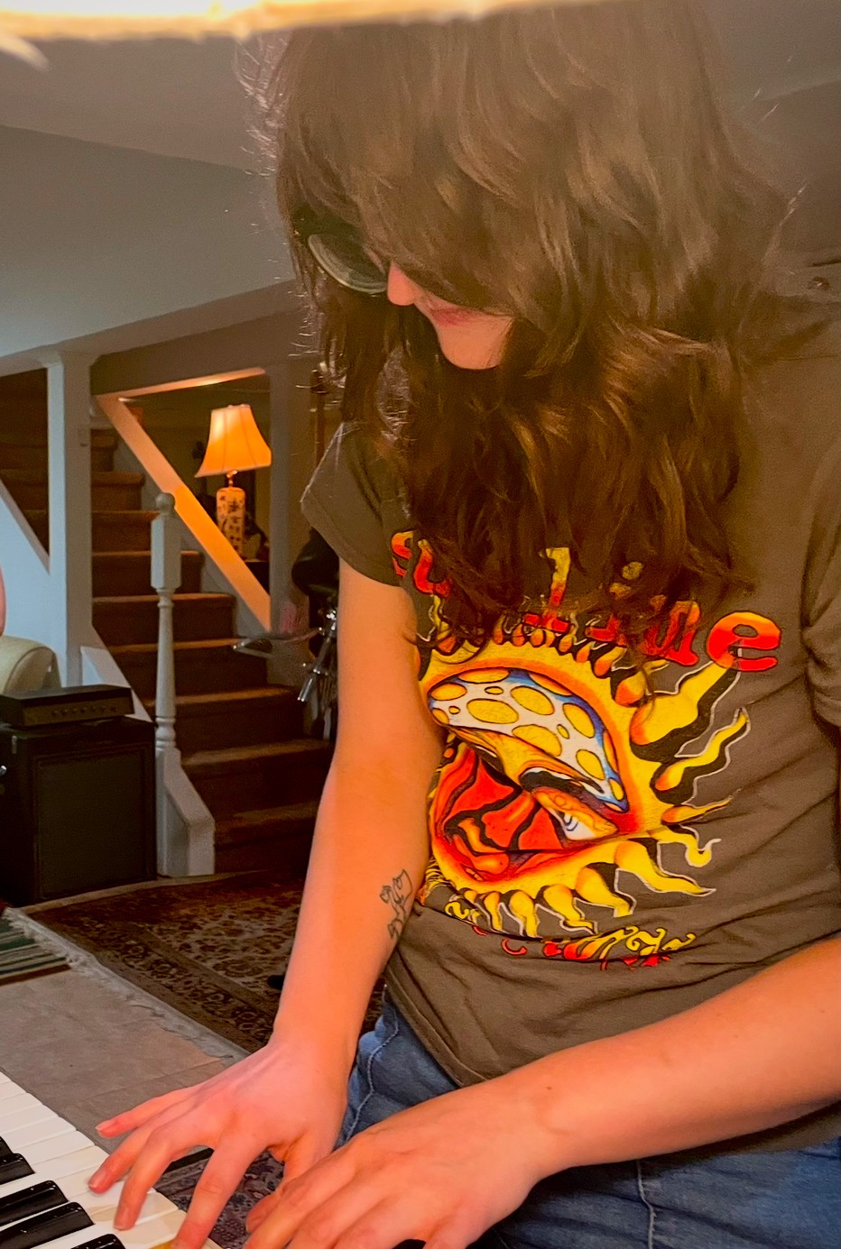 Person with long dark hair playing a keyboard, wearing a graphic t-shirt, in a home interior.