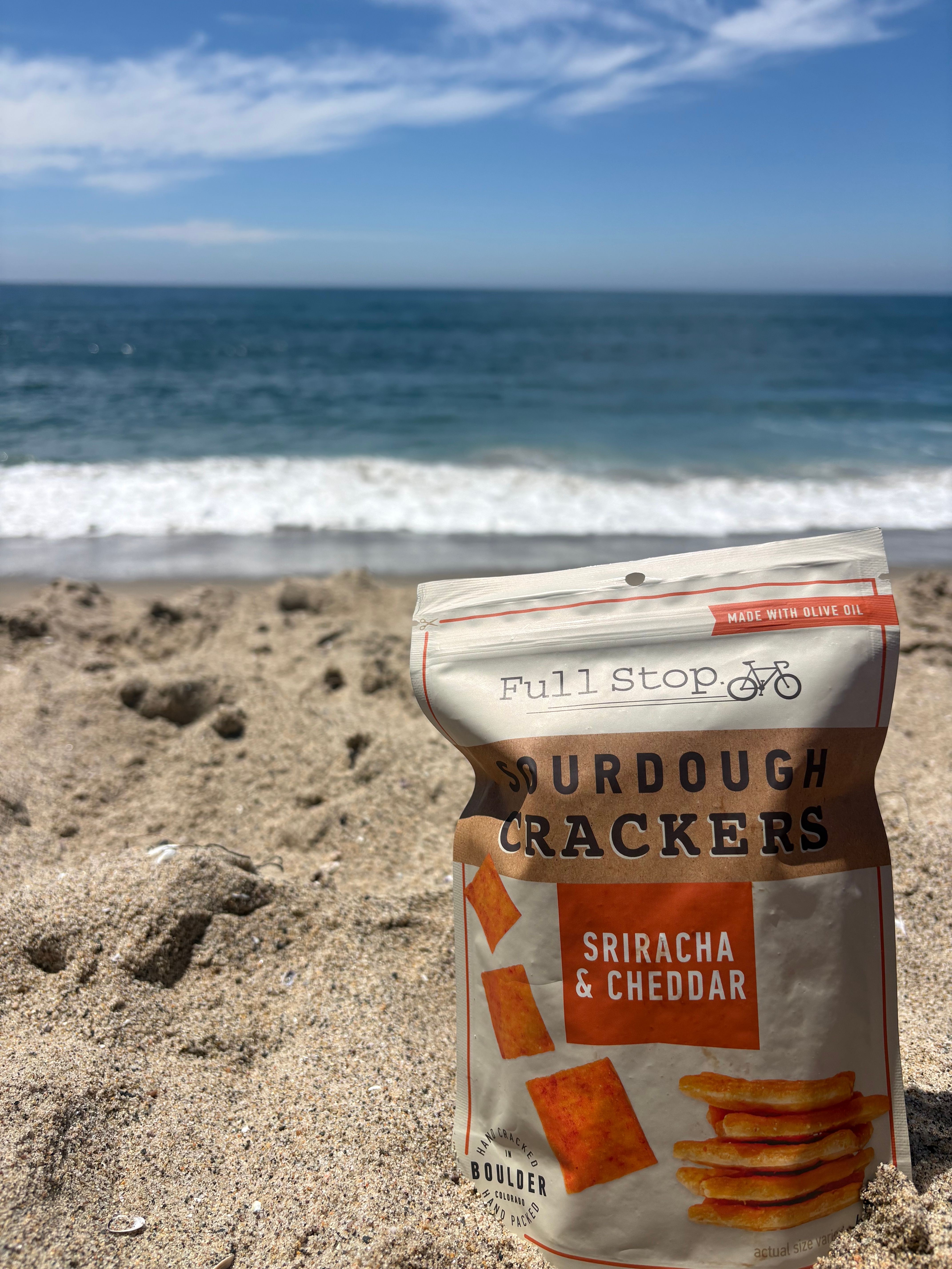 Bag of crunchy jerky snacks on sandy beach with ocean and sky in background.