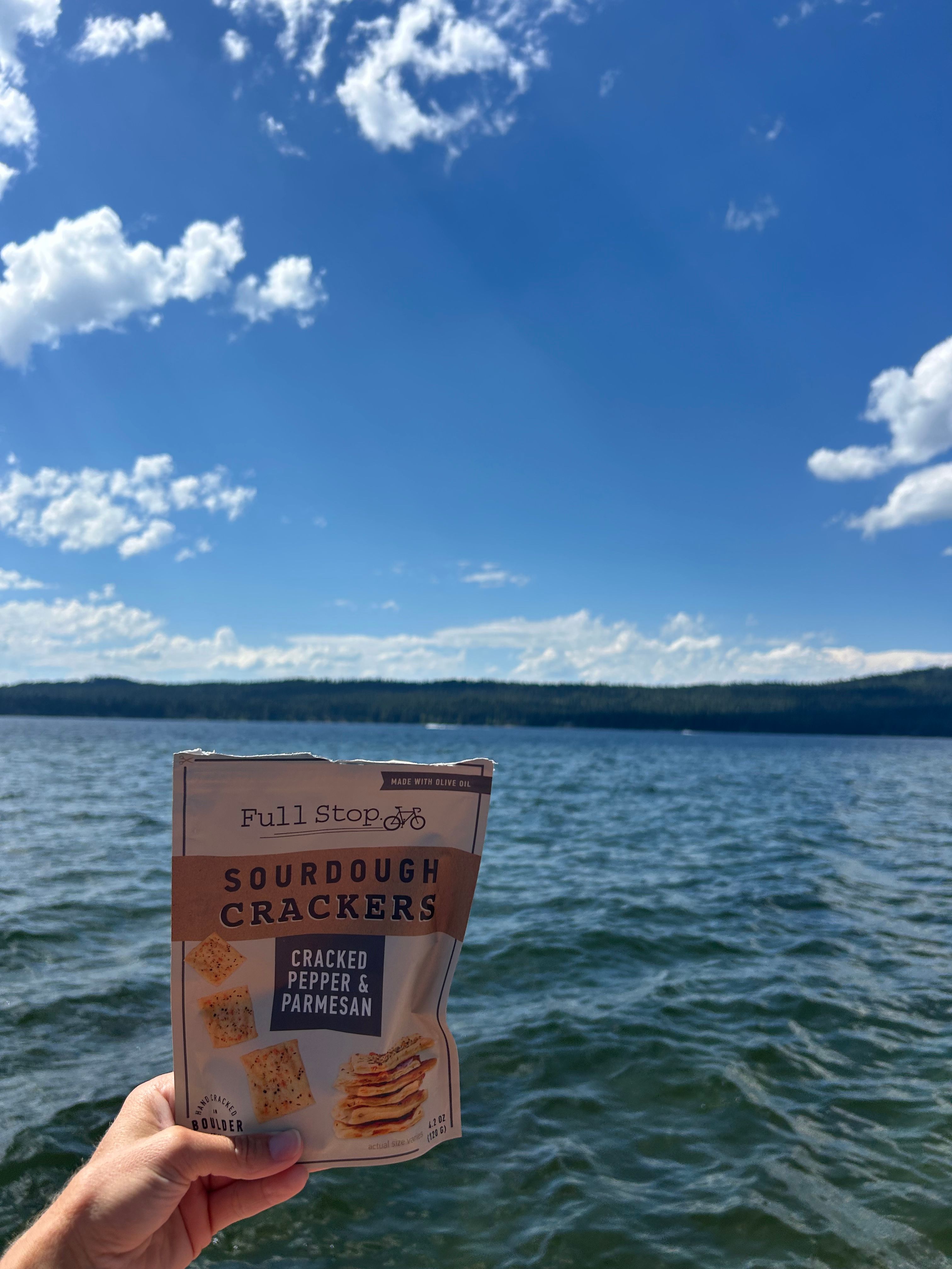 Person holding a book titled 'Surrounded by Cracker' near water with a blue sky and clouds.
