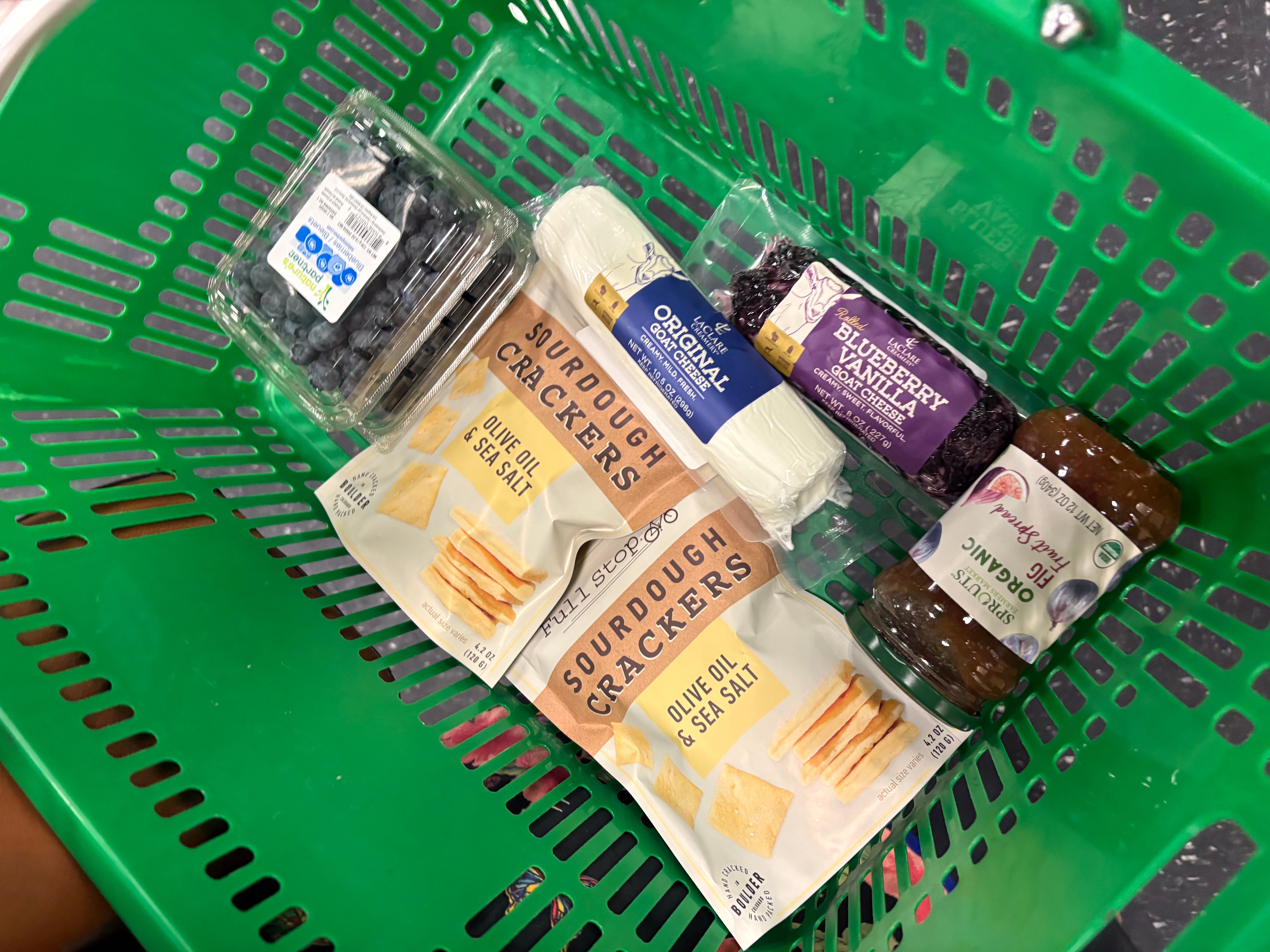 Green shopping basket containing packaged food items, including crackers, a jar, and other snack packages.