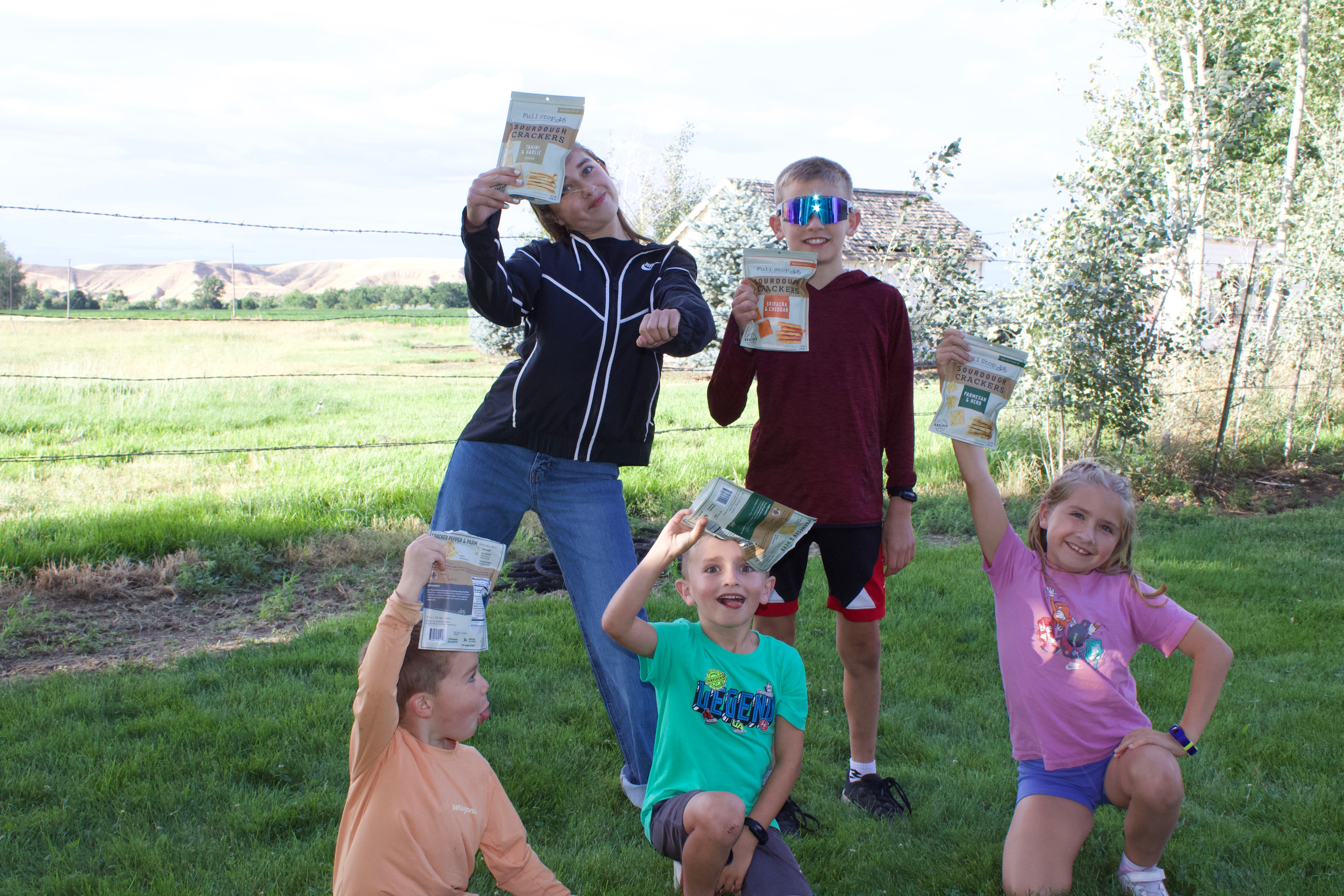 Five children outdoors holding magazines, smiling, and posing in a grassy area with trees and open field in background.