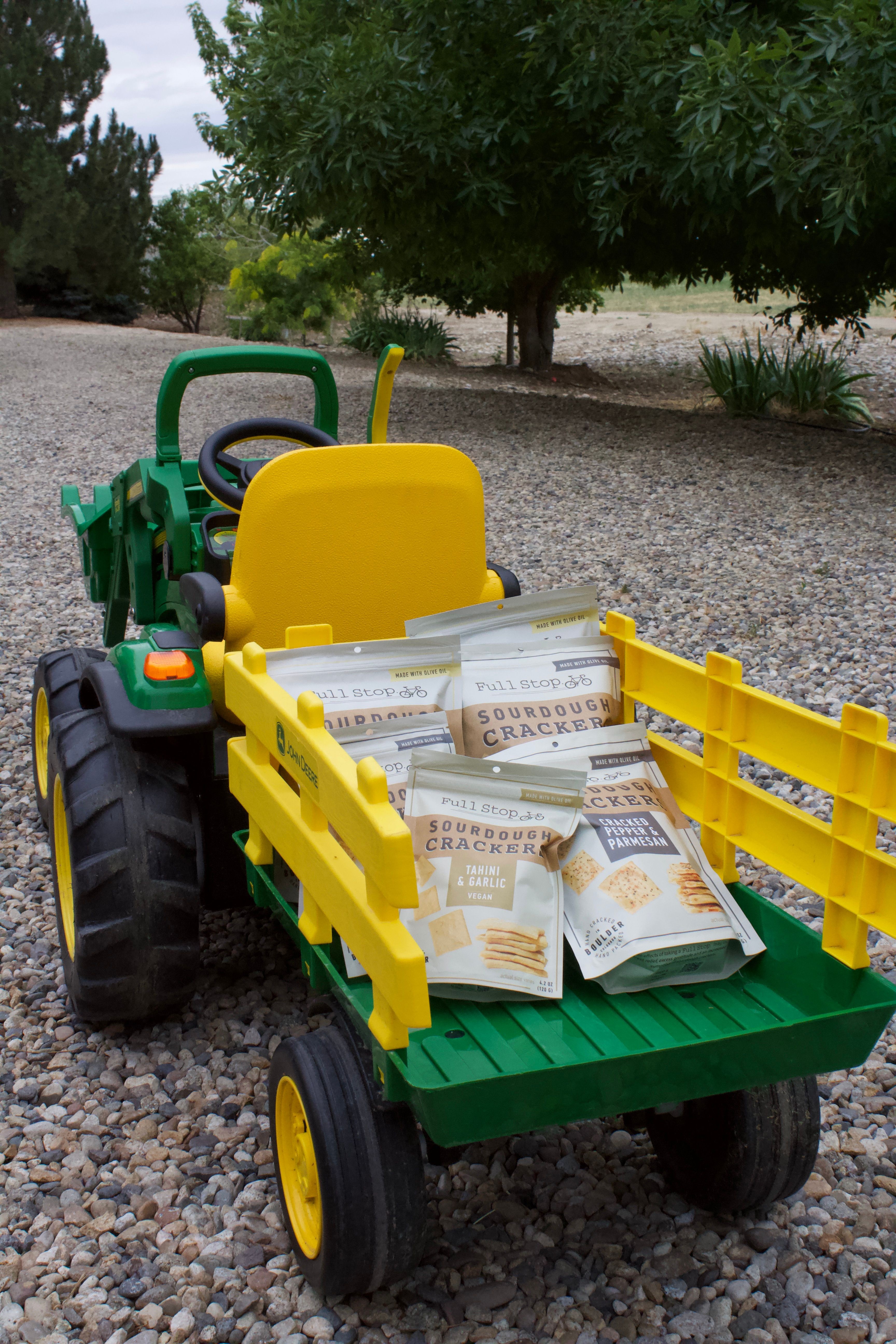 Toy green and yellow tractor with a yellow seat and black wheels on gravel, with trees in the background.