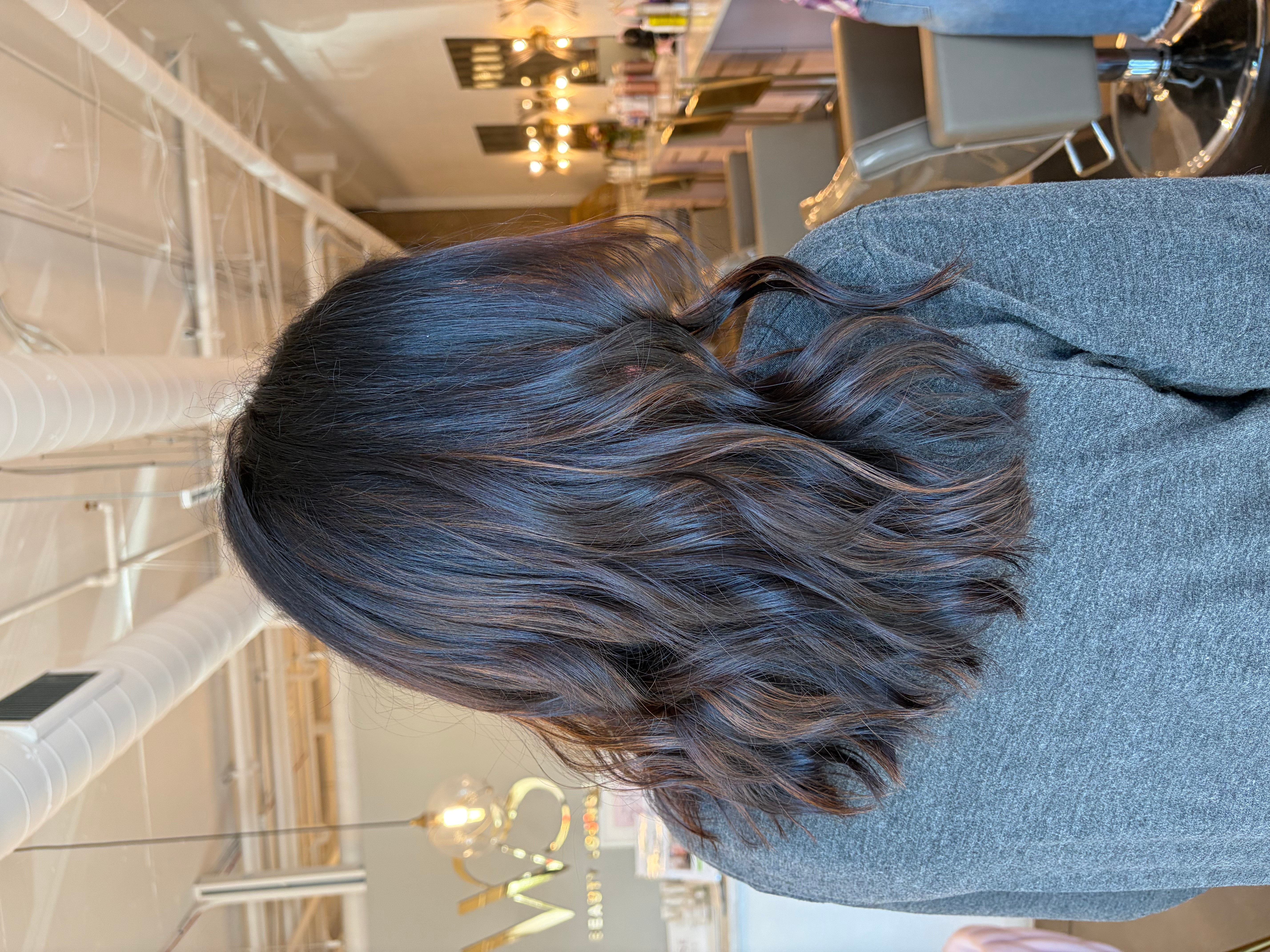 Person with shoulder-length wavy hair wearing a gray top, viewed from behind in an indoor setting.