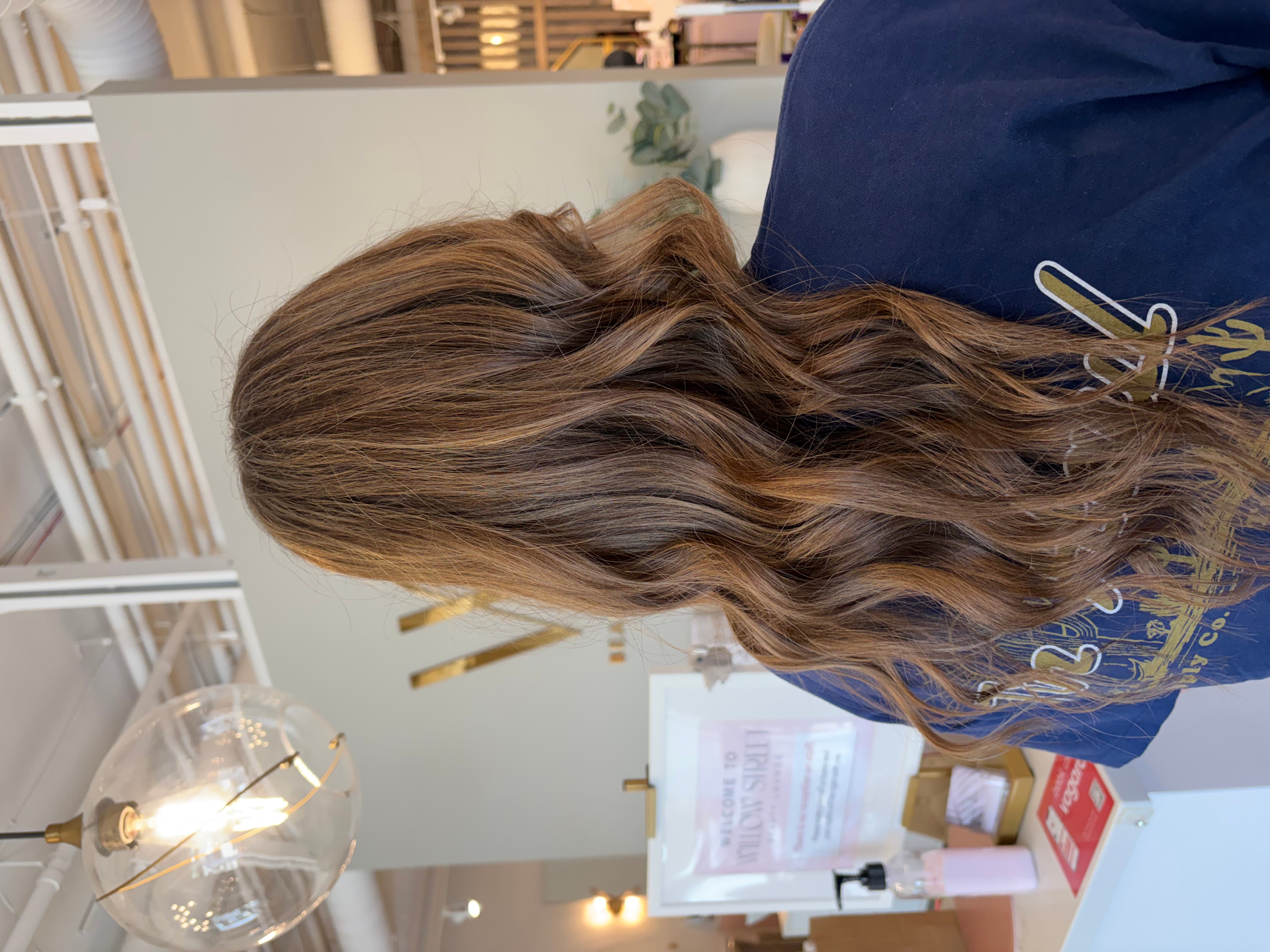 Person with long, wavy brown hair viewed from the side, wearing a dark blue top, in an indoor setting.