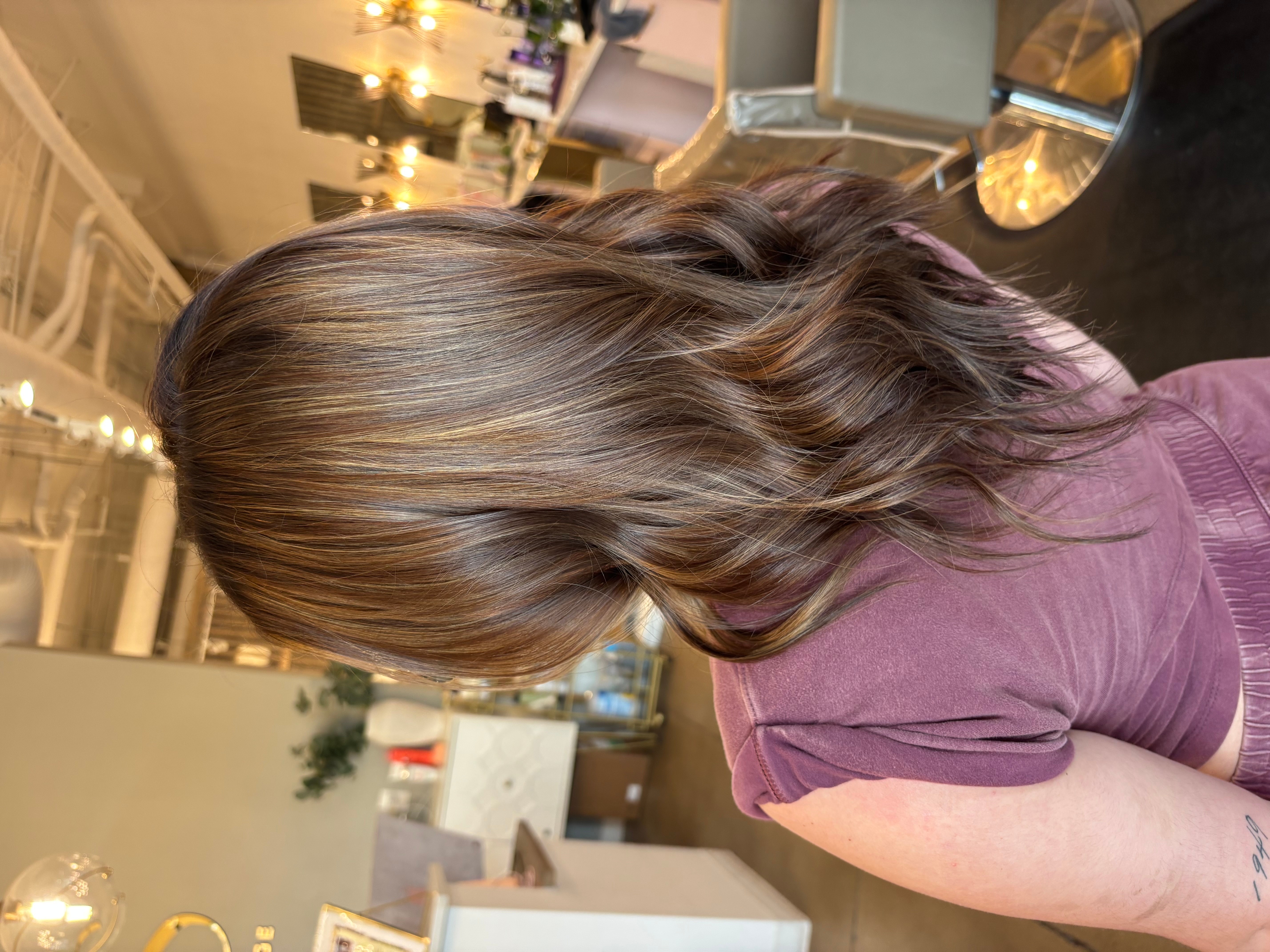 Side view of a woman with wavy brown hair wearing a purple shirt in a room with furniture and decorations.