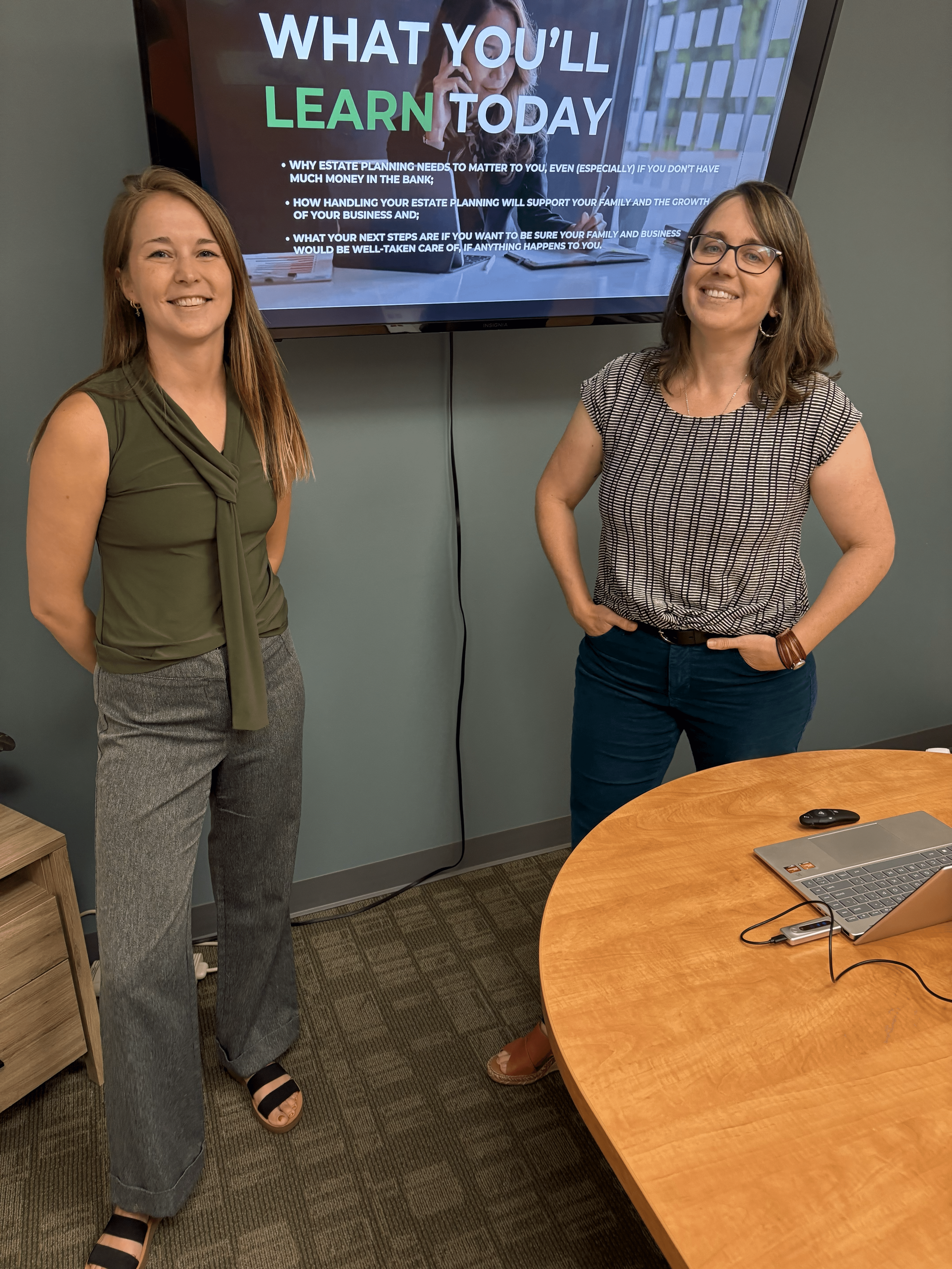 Two women stand in front of a screen displaying text, smiling, in a room with a wooden table and a laptop.