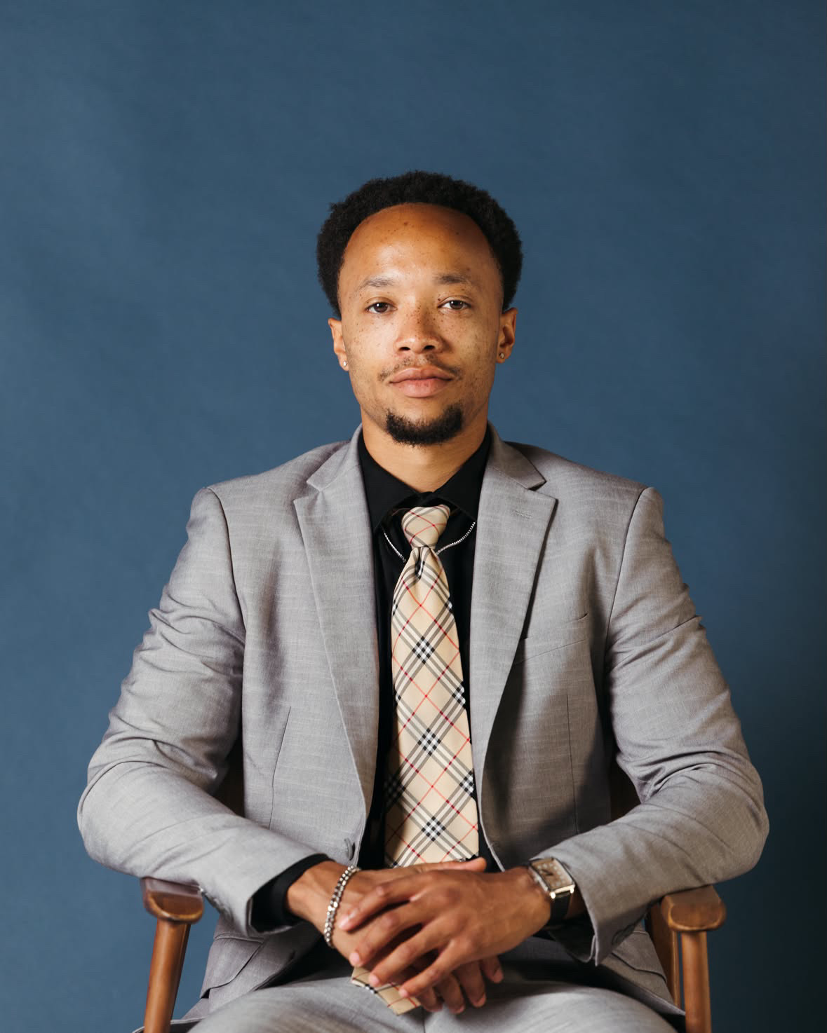 Man in a gray suit and plaid tie sitting on a chair against a blue background.
