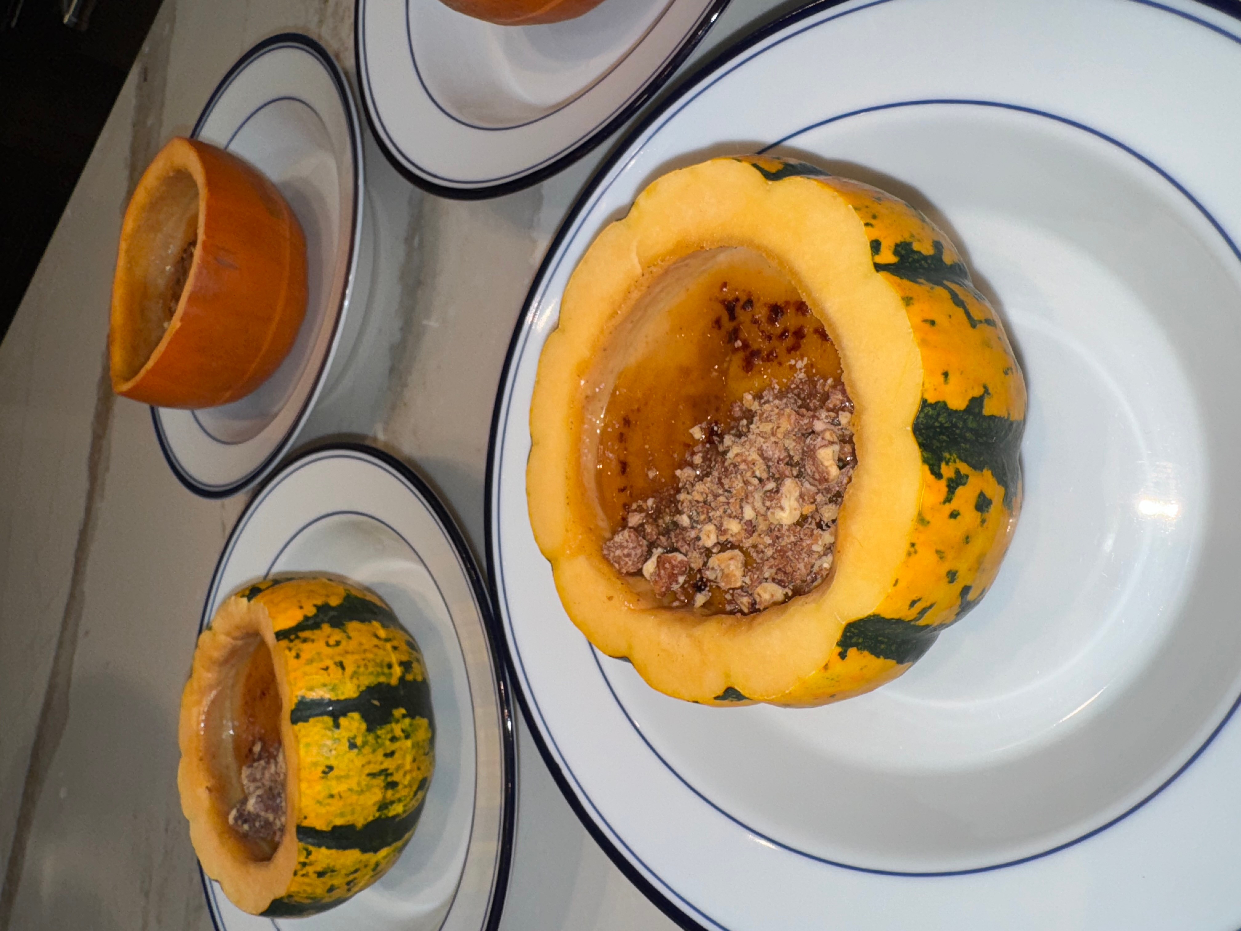 Gourds filled with a mixture of ingredients, placed on white plates with blue lines, and a halved orange on a table.