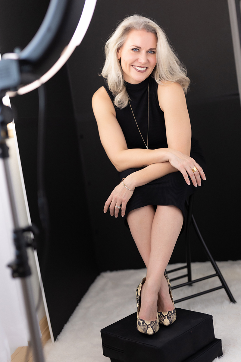Woman with blonde hair smiling, sitting on a stool with crossed legs, in a black dress, in a studio setting.
