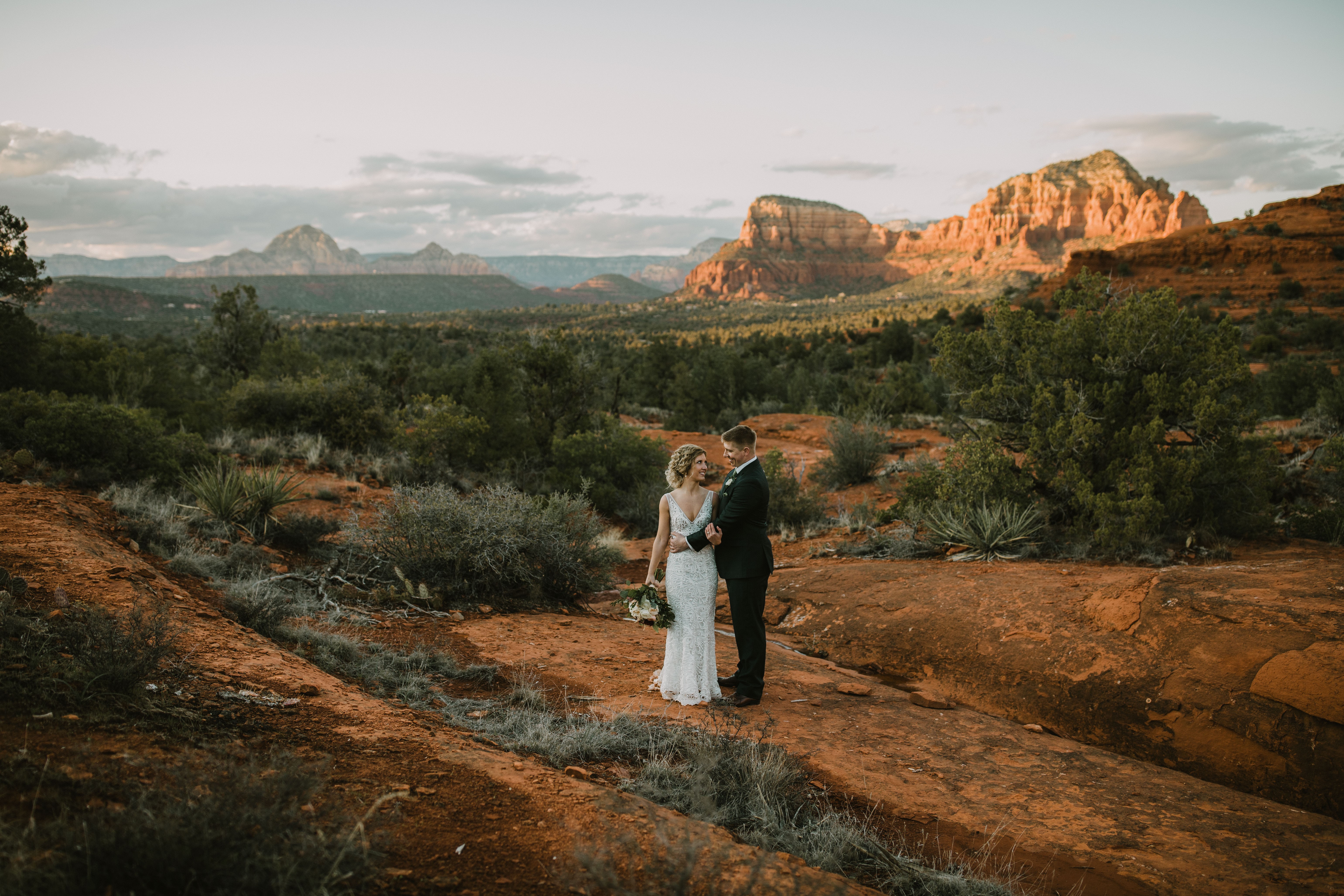 A couple in wedding attire standing close together in a desert landscape with mountains in the background.