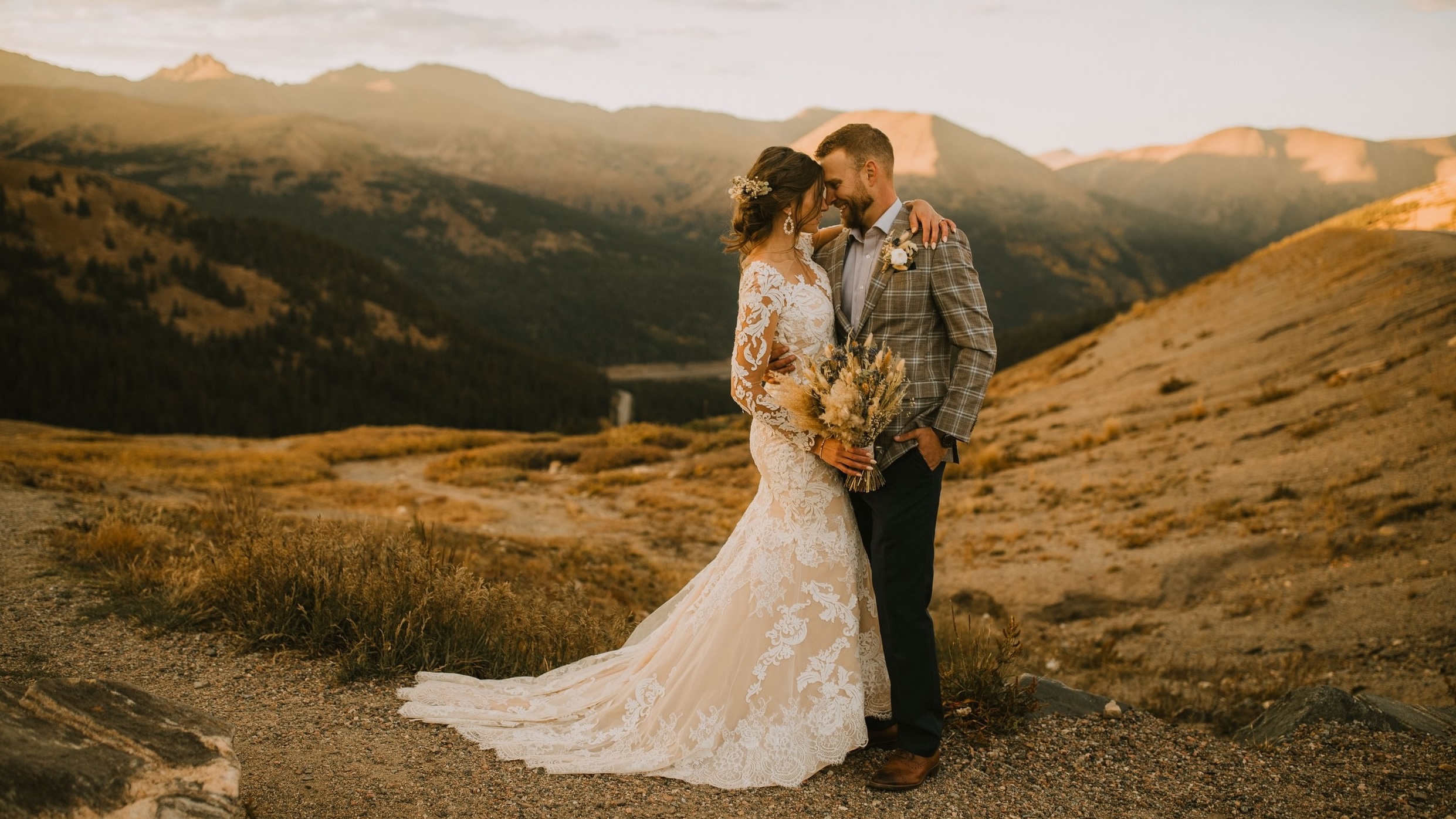 A bride and groom stand close together outdoors with mountains in the background during sunset.