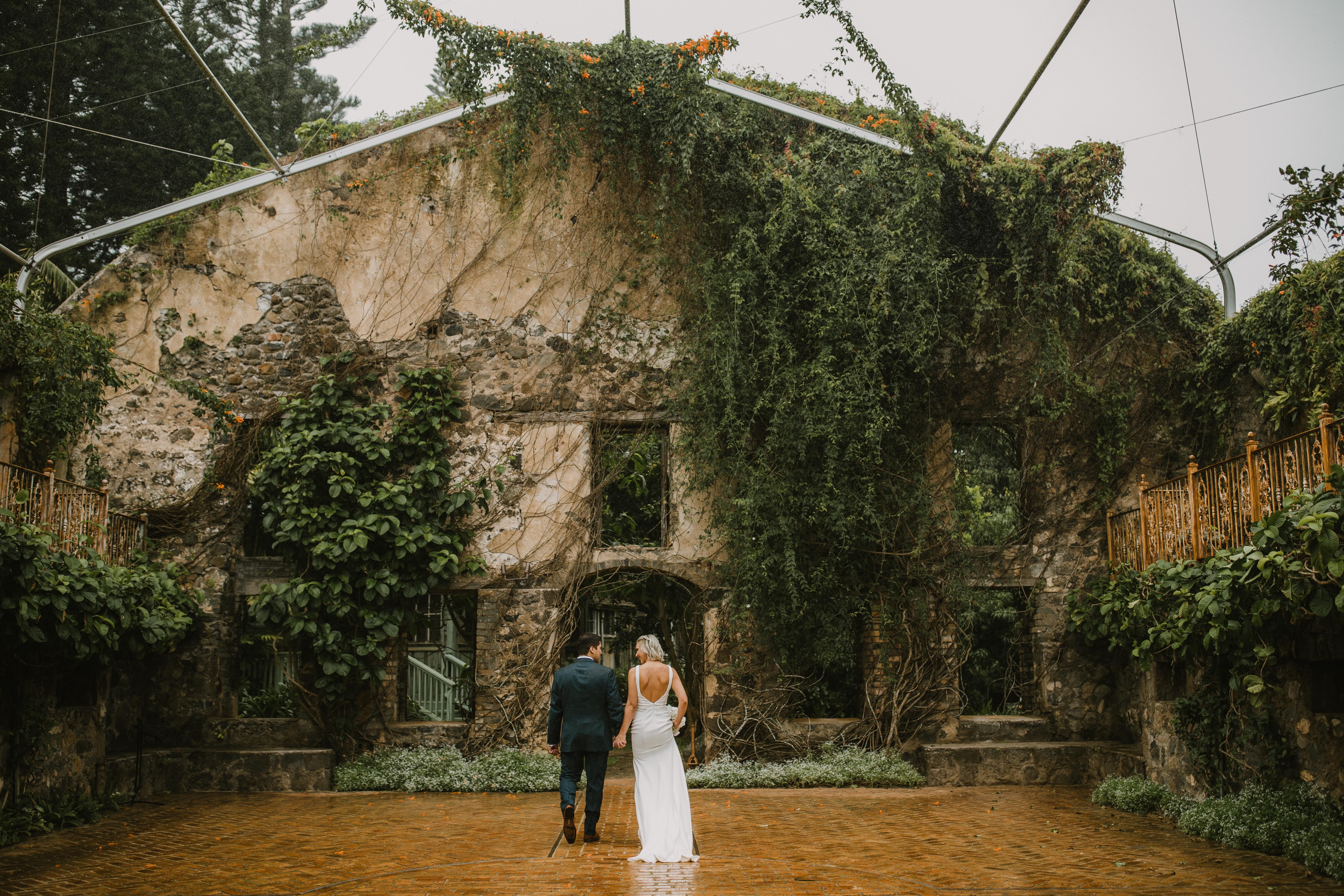 A couple in wedding attire standing in front of a stone building covered with greenery and vines, under a glass ceiling.