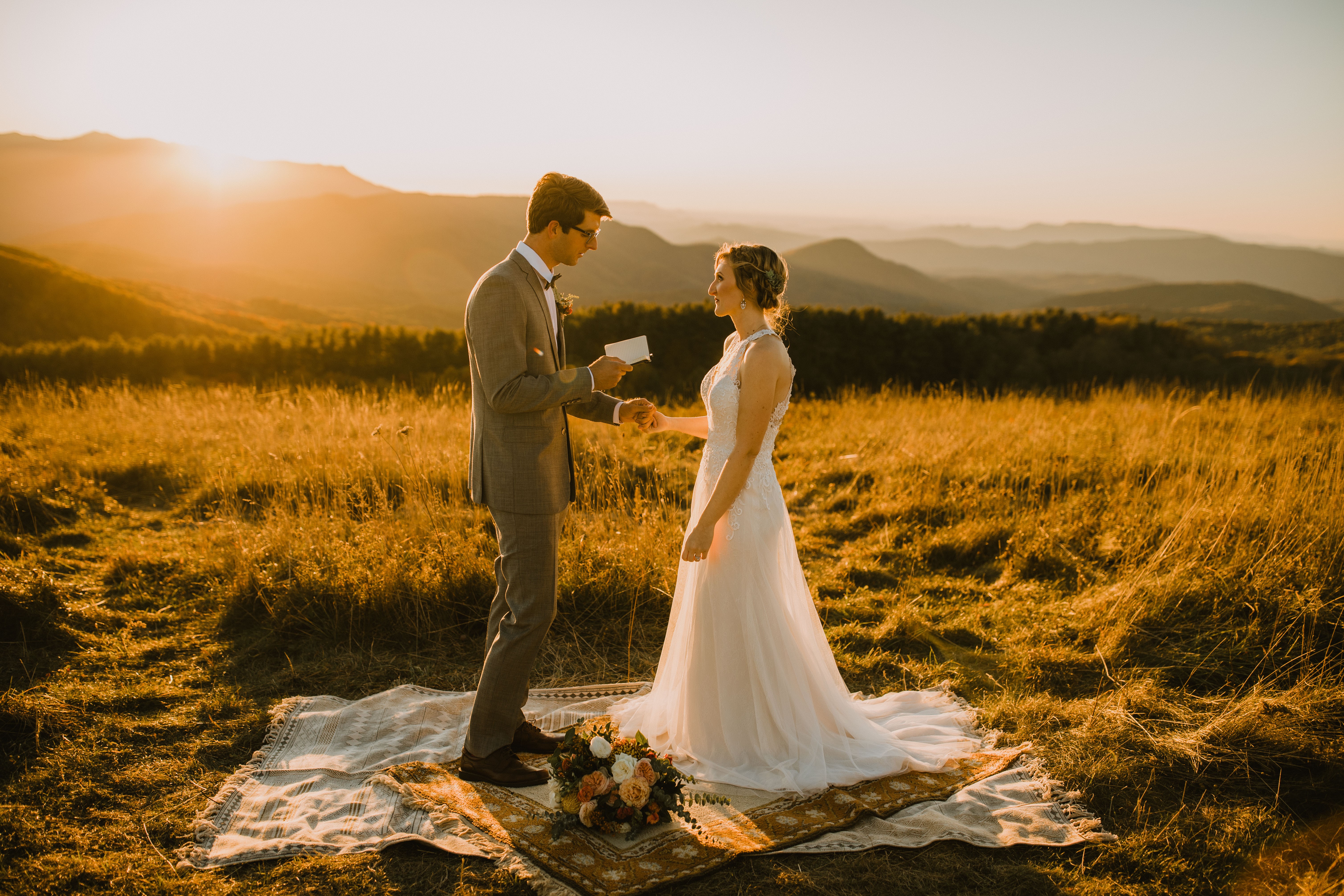 A couple in wedding attire standing on a blanket in a field during sunset, exchanging rings with mountains in the background.