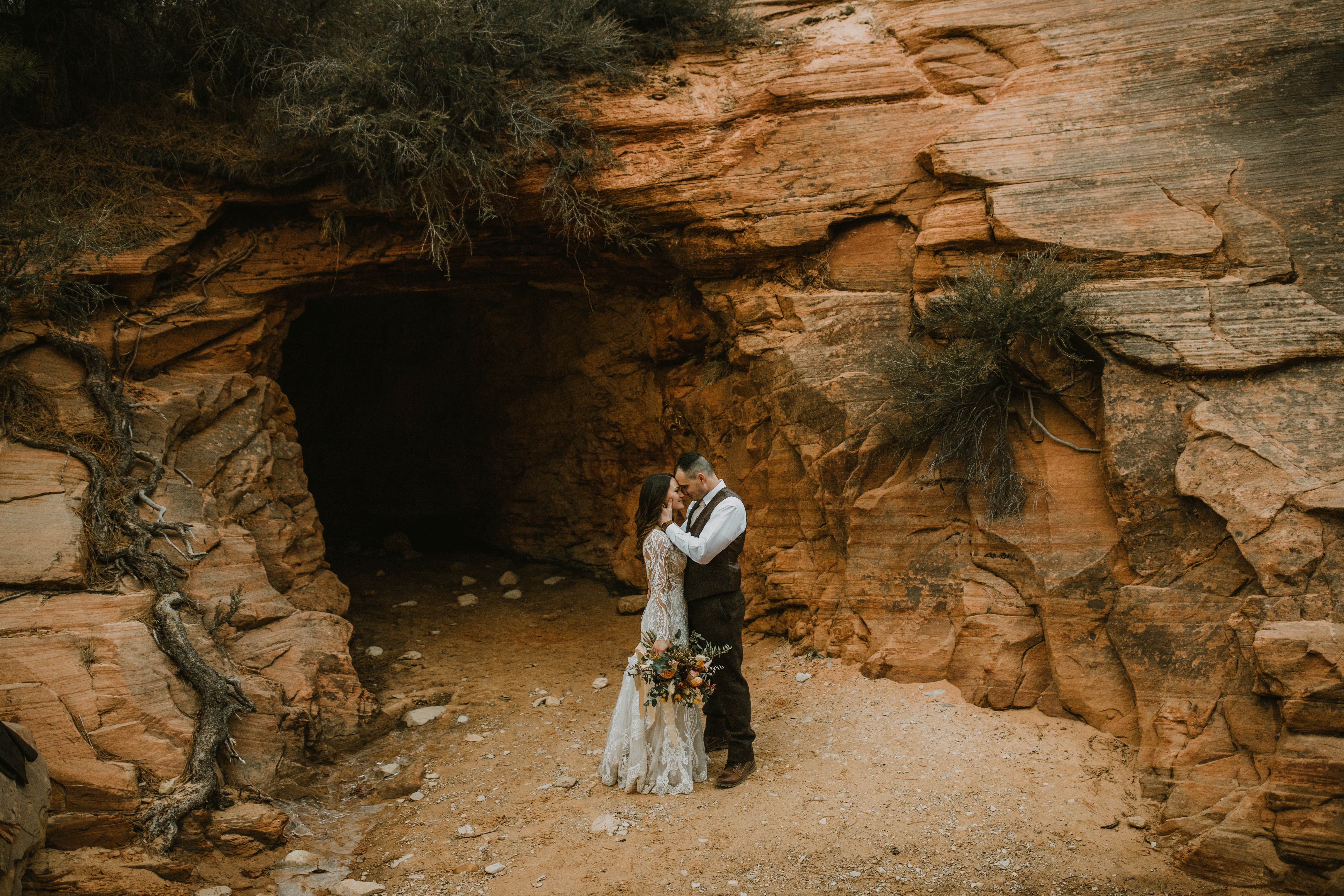 A couple stands close together in front of a rocky cave entrance, holding a bouquet of flowers, outdoors.