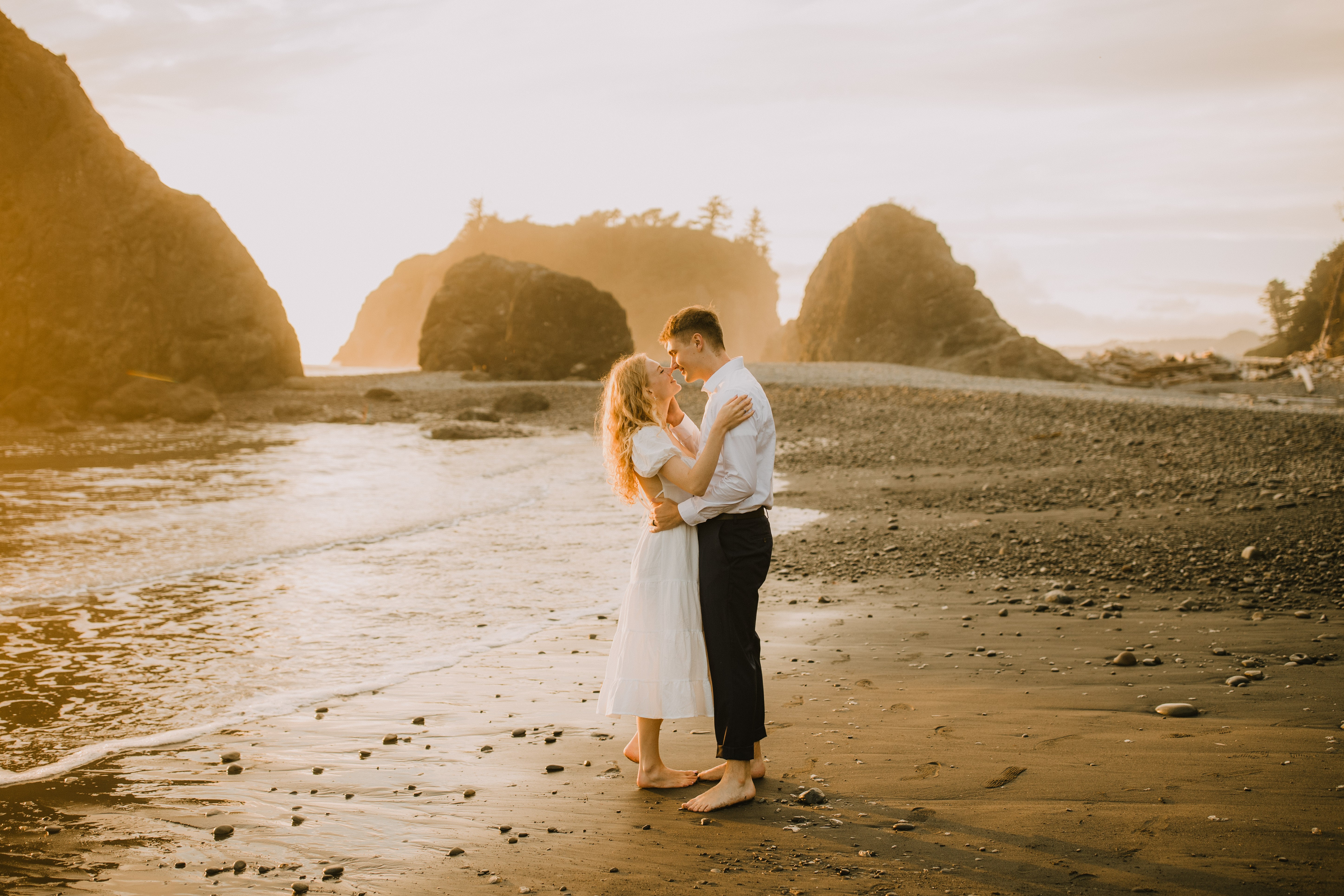 A couple stands on a beach, embracing with large rock formations in the background during sunset.