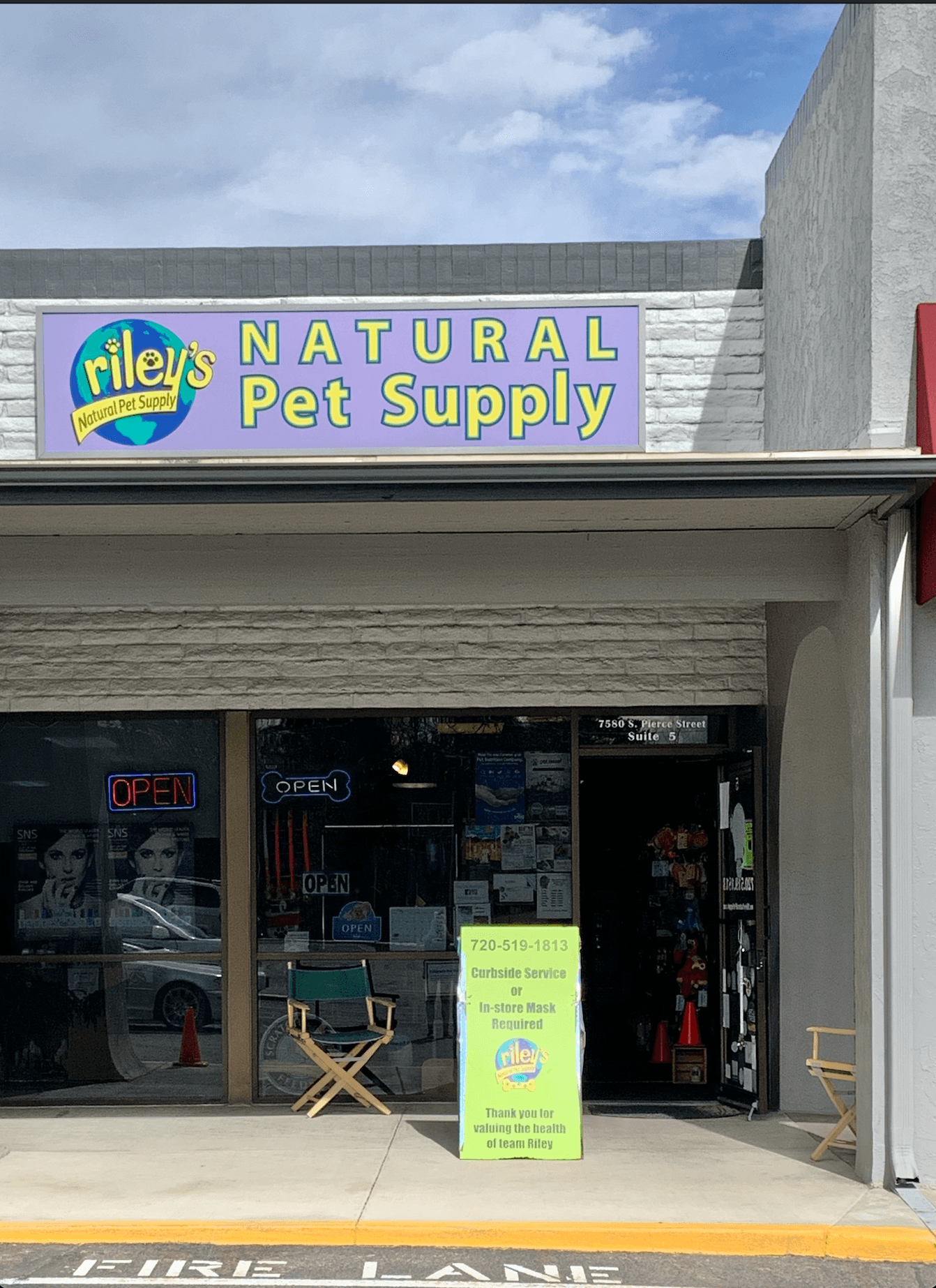 Storefront with sign reading 'Riley's Natural Pet Supply' and an open door, outdoor chairs, and a yellow sign outside.