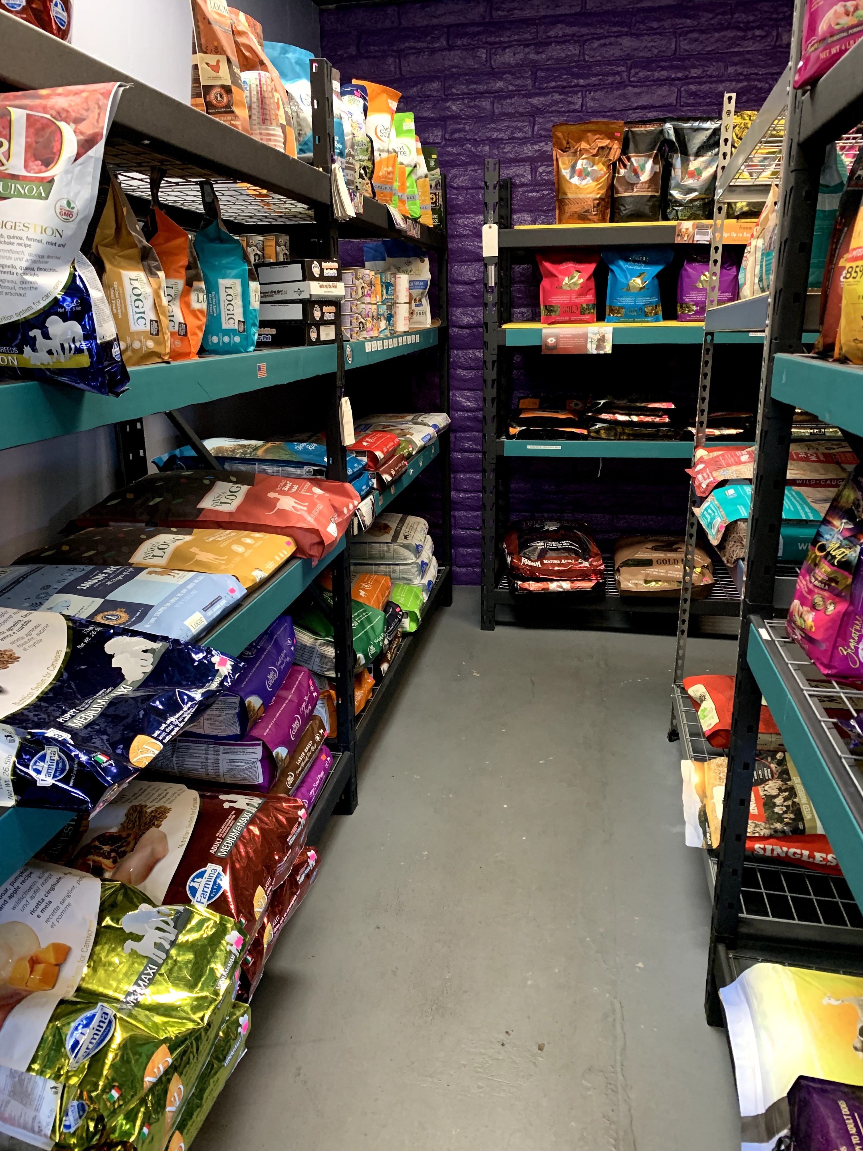 Shelves stocked with various bags of pet food in a store aisle, with a purple brick wall in the background.