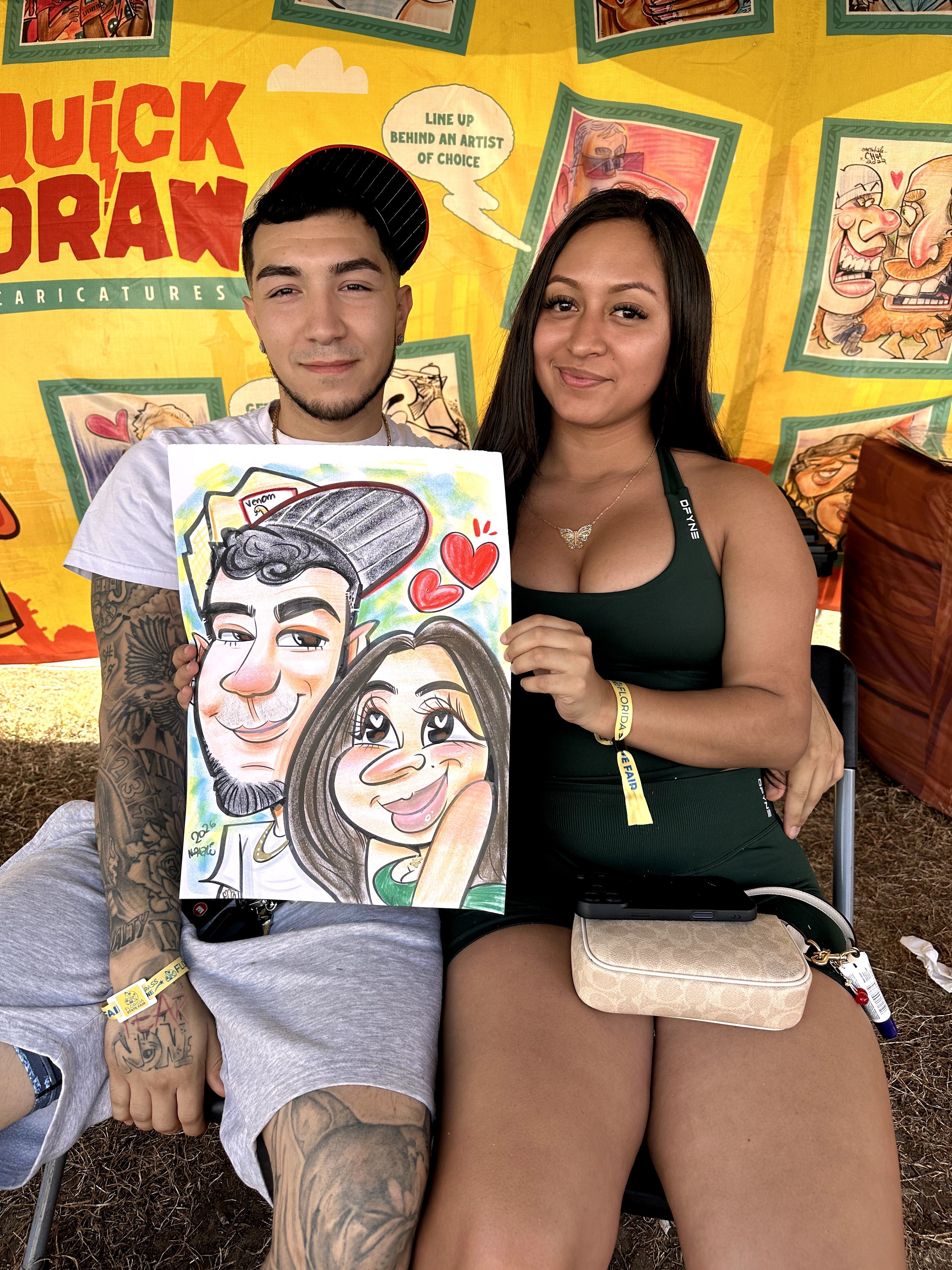 A young man and woman sit together, holding a caricature drawing of themselves with hearts, in front of a colorful background.
