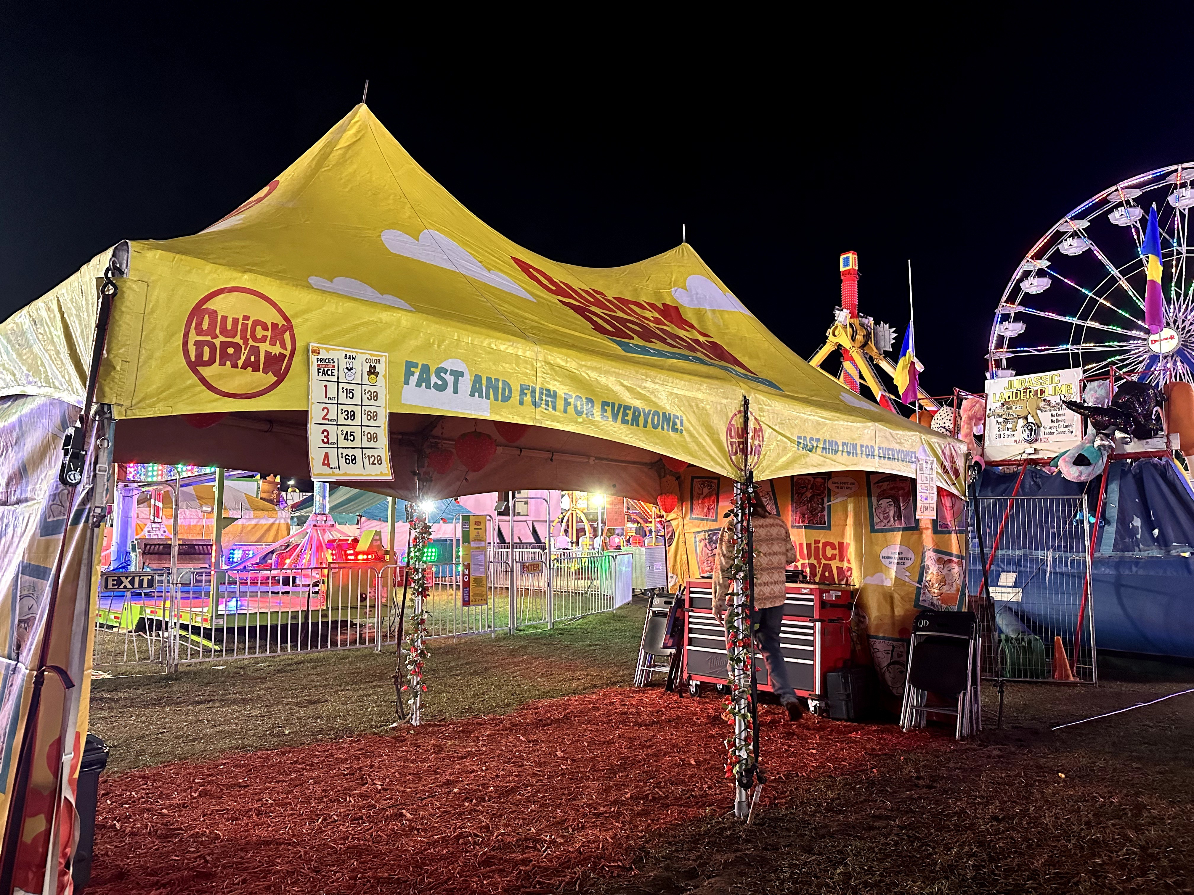 Nighttime carnival with a yellow tent, rides, and colorful lights, surrounded by a fence and a dirt ground.