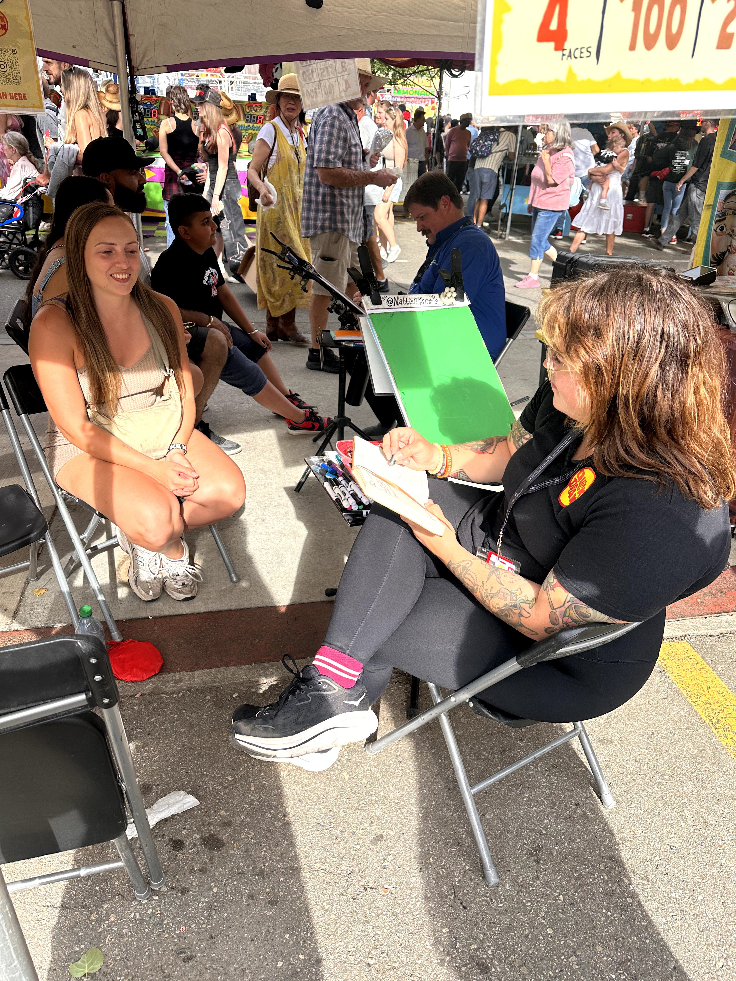 Two women sitting outdoors at a market or fair, one with a clipboard and the other smiling, with people and tents in background.