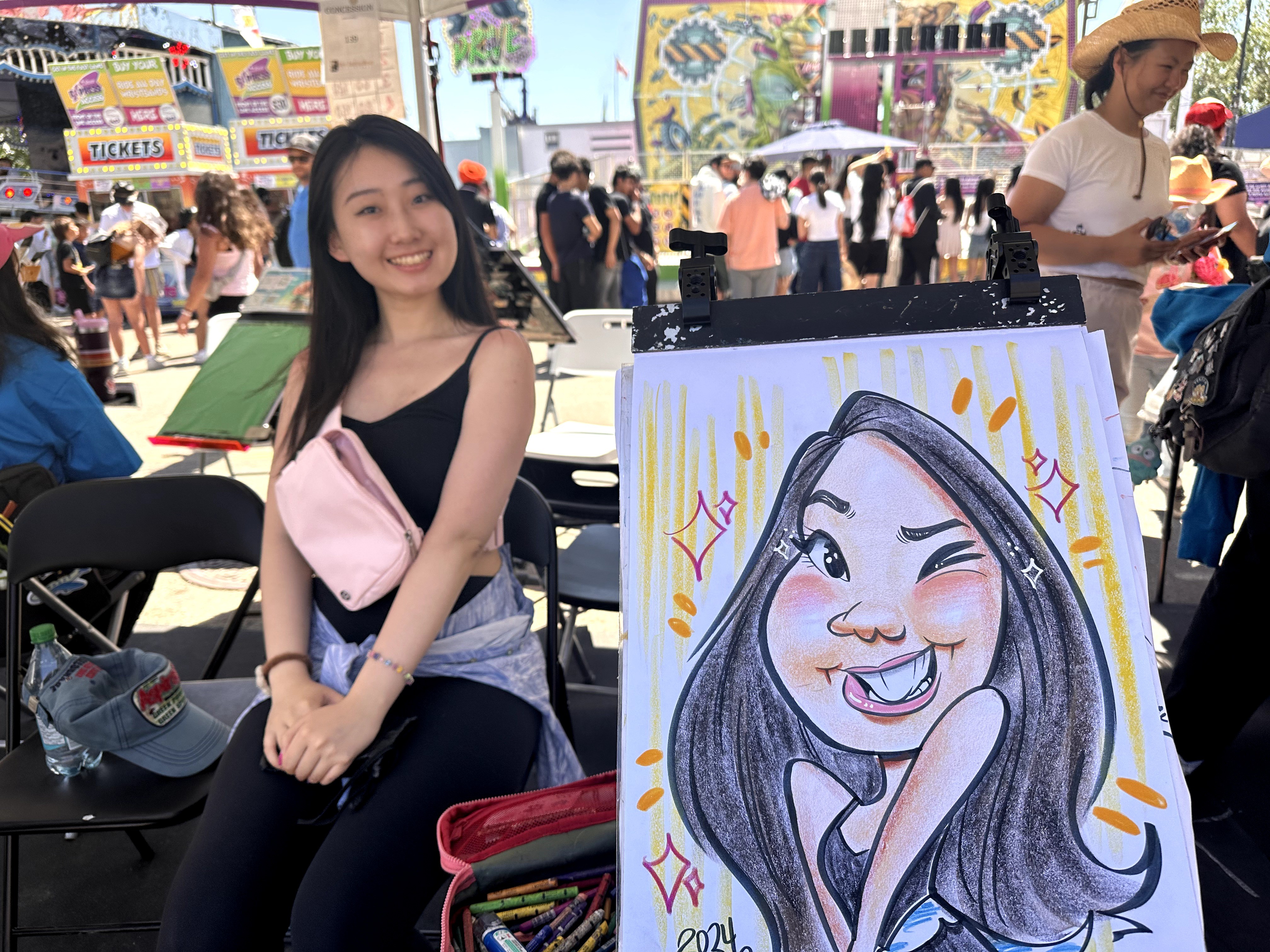 Young woman smiling at an outdoor event, standing next to a colorful caricature drawing on an easel, with a crowd in the background.