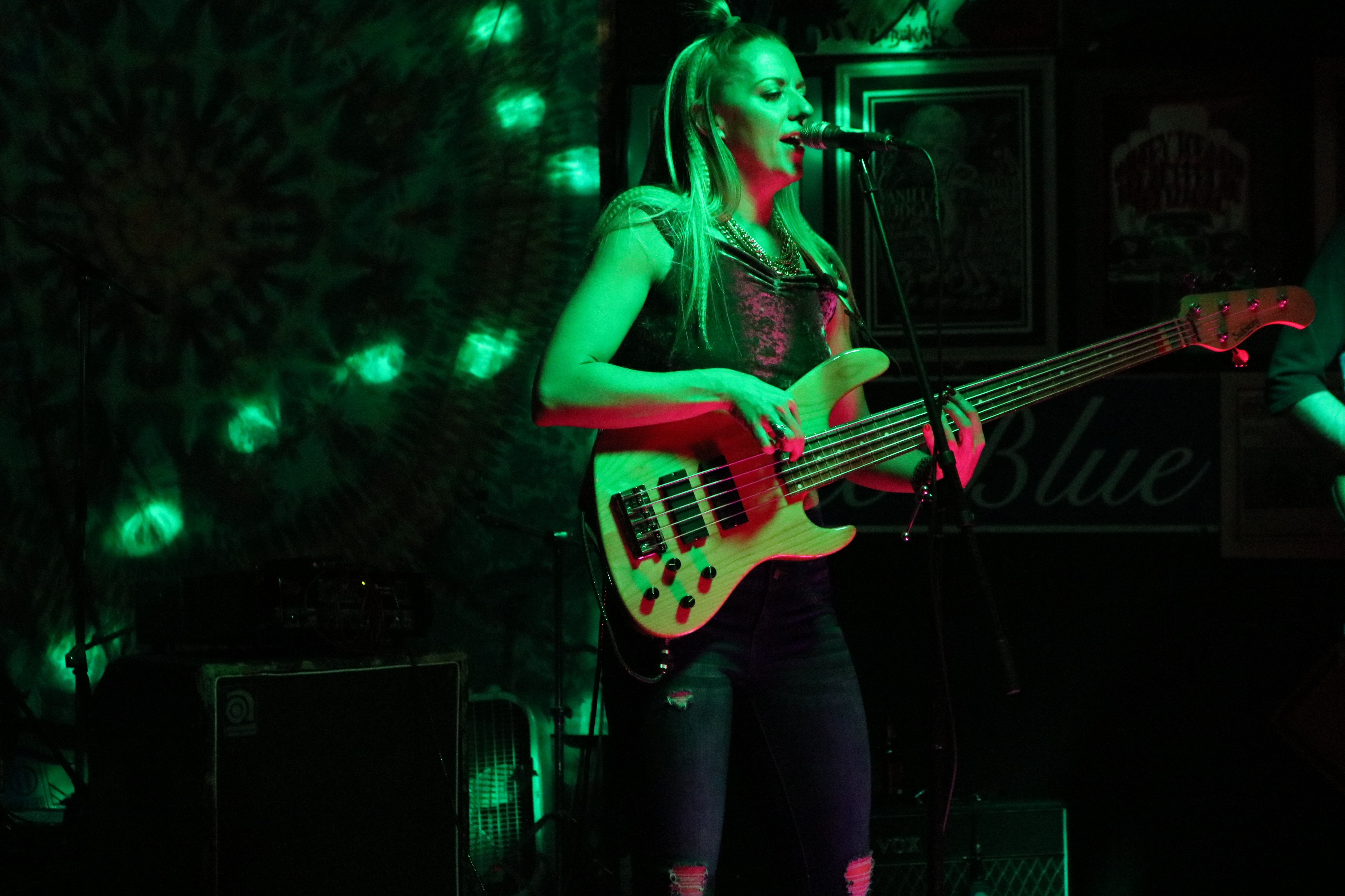 A woman playing a red bass guitar on stage with green lighting, singing into a microphone.