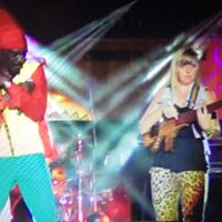 Two women performing on stage with colorful lighting and musical instruments, one wearing a red jacket and the other leopard print pants.