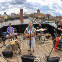 Musicians perform on a rooftop with city buildings in the background, playing instruments and singing. Cloudy sky overhead.
