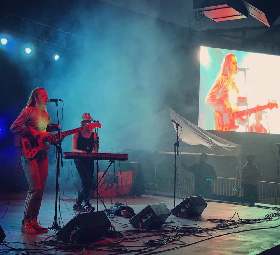 Two musicians perform on stage with a large screen showing a close-up of a guitarist, stage lights, and fog effects.