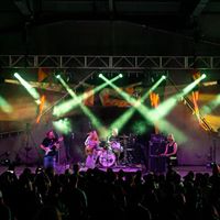 Band performs on stage with colorful lights, audience watching, in a concert setting.