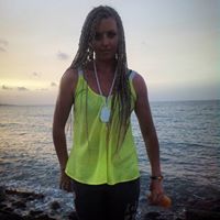 Woman standing by the water at sunset, wearing a bright yellow top and dark pants, with long braided hair.