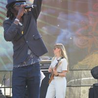 A person in a dark suit holding a microphone, and a woman in white uniform standing outdoors.