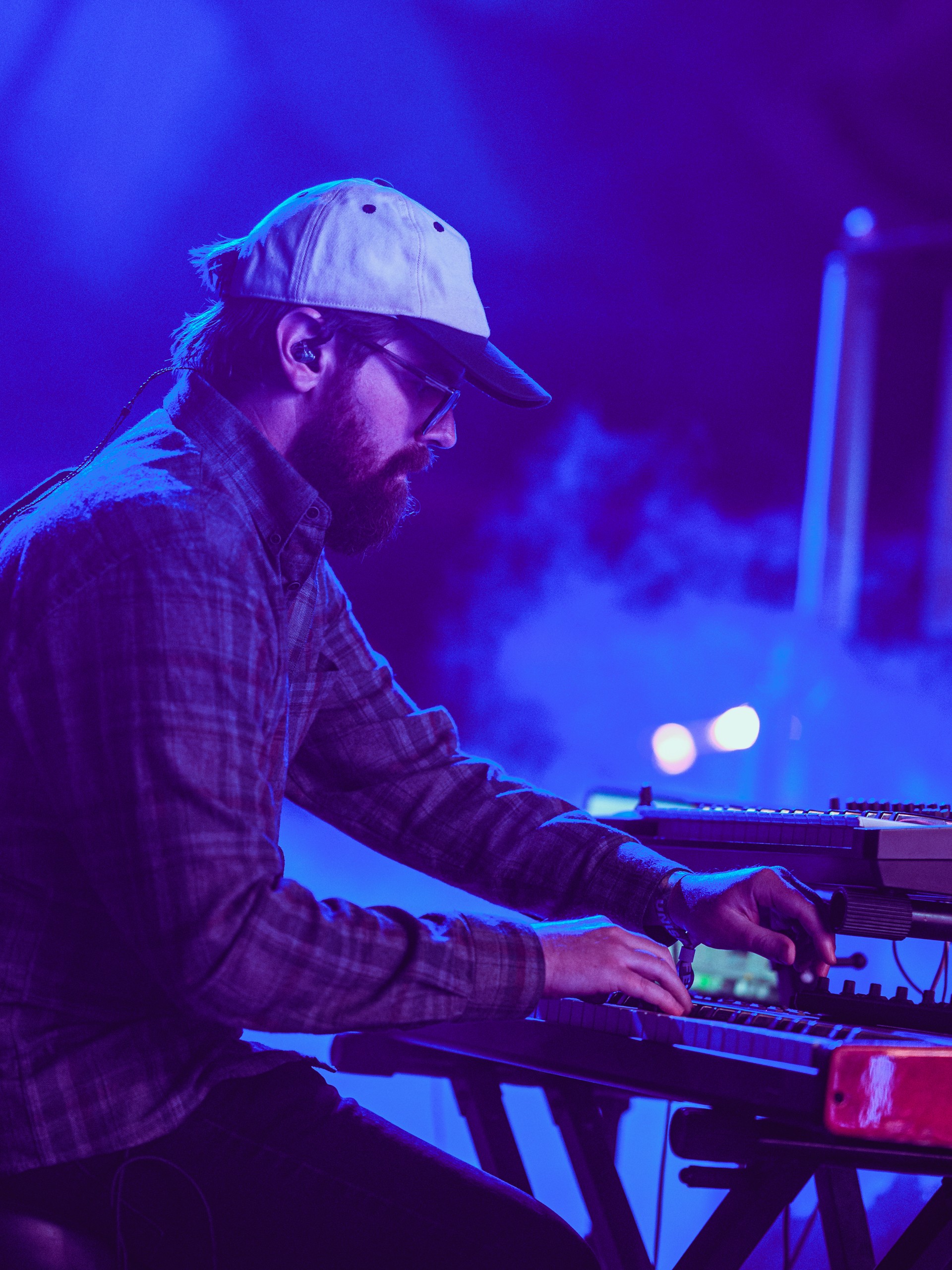 Person playing keyboard on stage with blue and purple lighting, wearing a cap and glasses, focused on music.