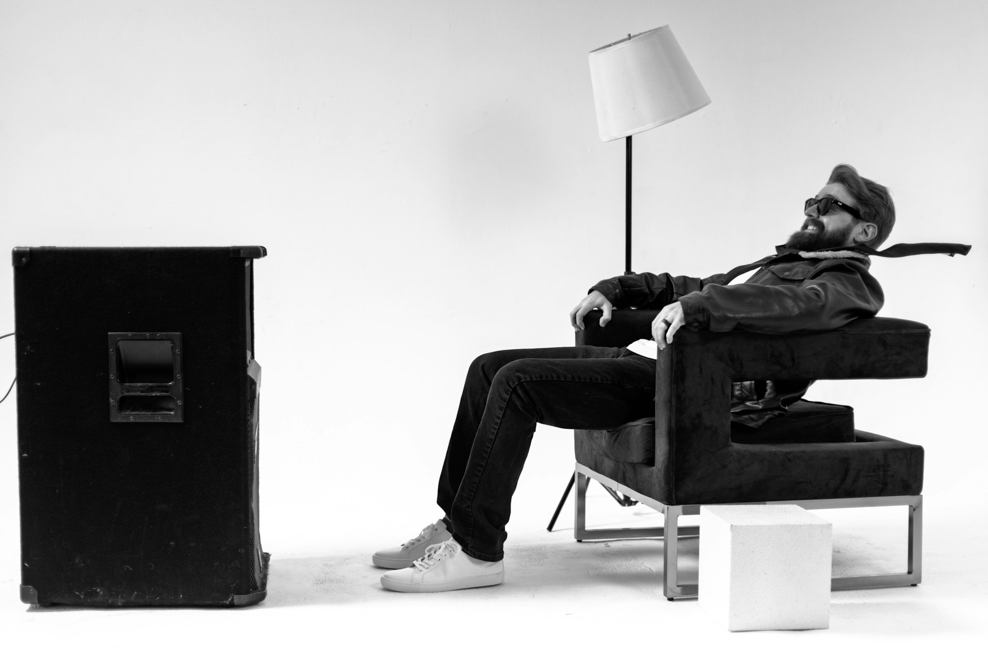 Man with a beard and glasses relaxes on a sofa with a lamp behind him, facing a cabinet, in a minimal room.