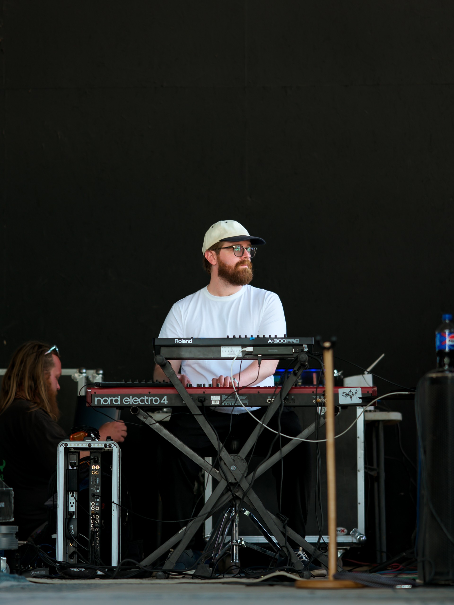 Person with glasses and a white cap standing behind a keyboard and electronic equipment, black background.