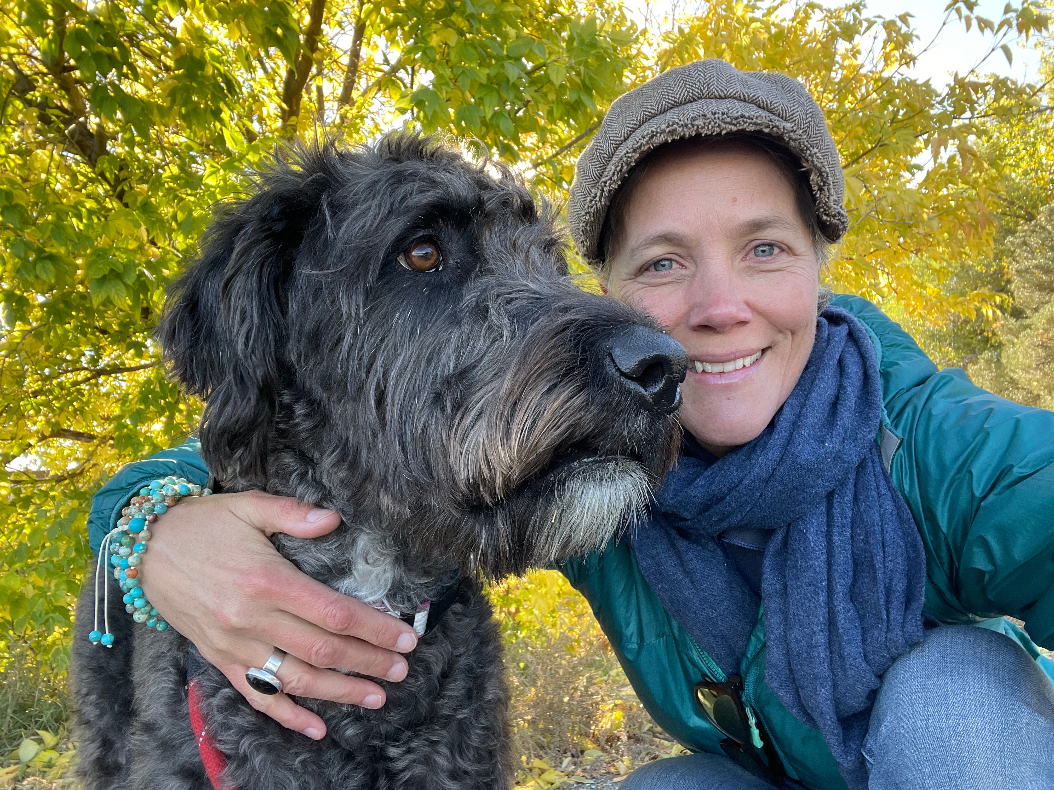 Woman smiling outdoors with a large, dark, shaggy dog, surrounded by green and yellow autumn leaves.