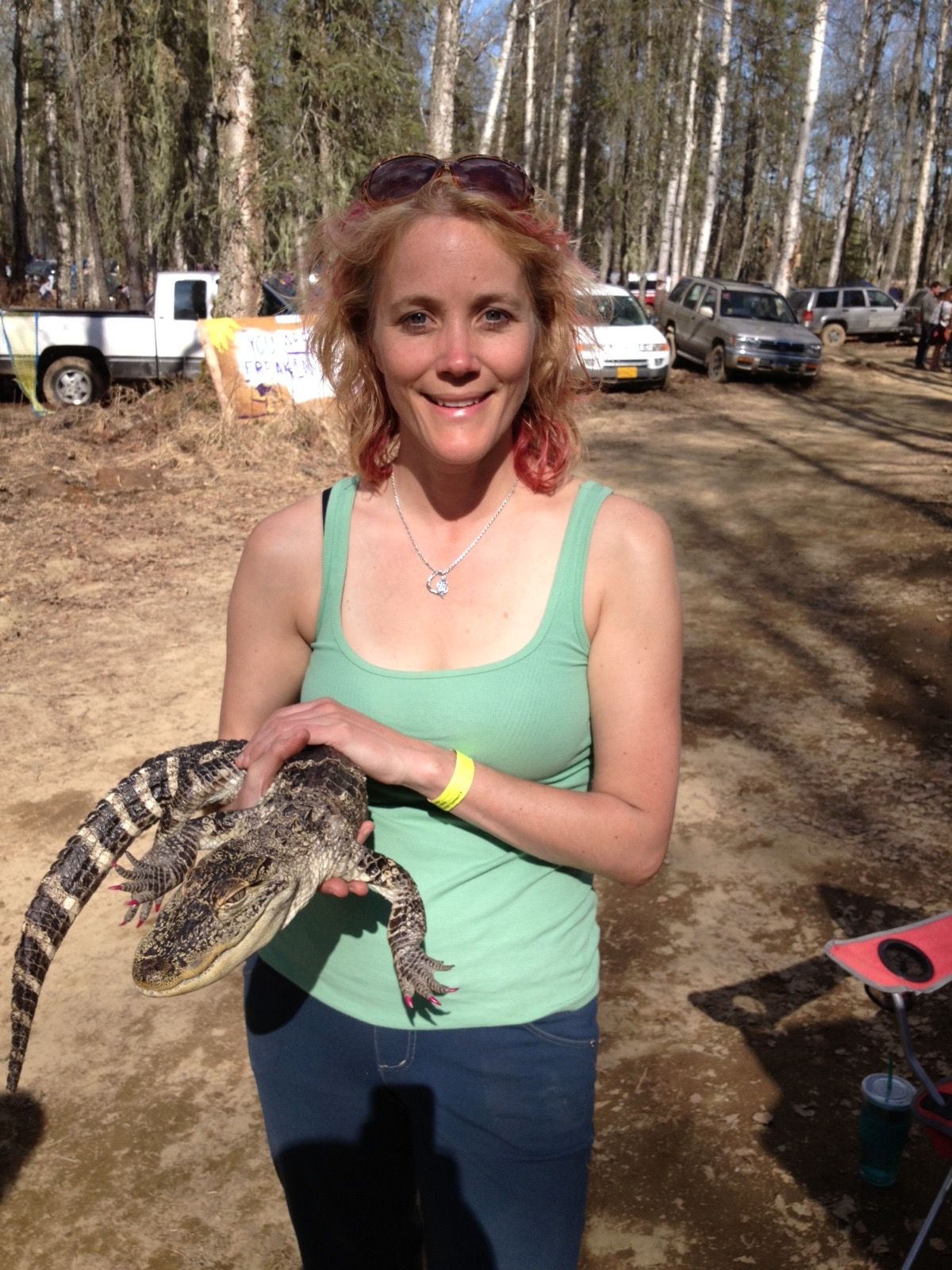 Woman holding a small alligator outdoors in a wooded area, smiling, wearing a green tank top and dark pants.