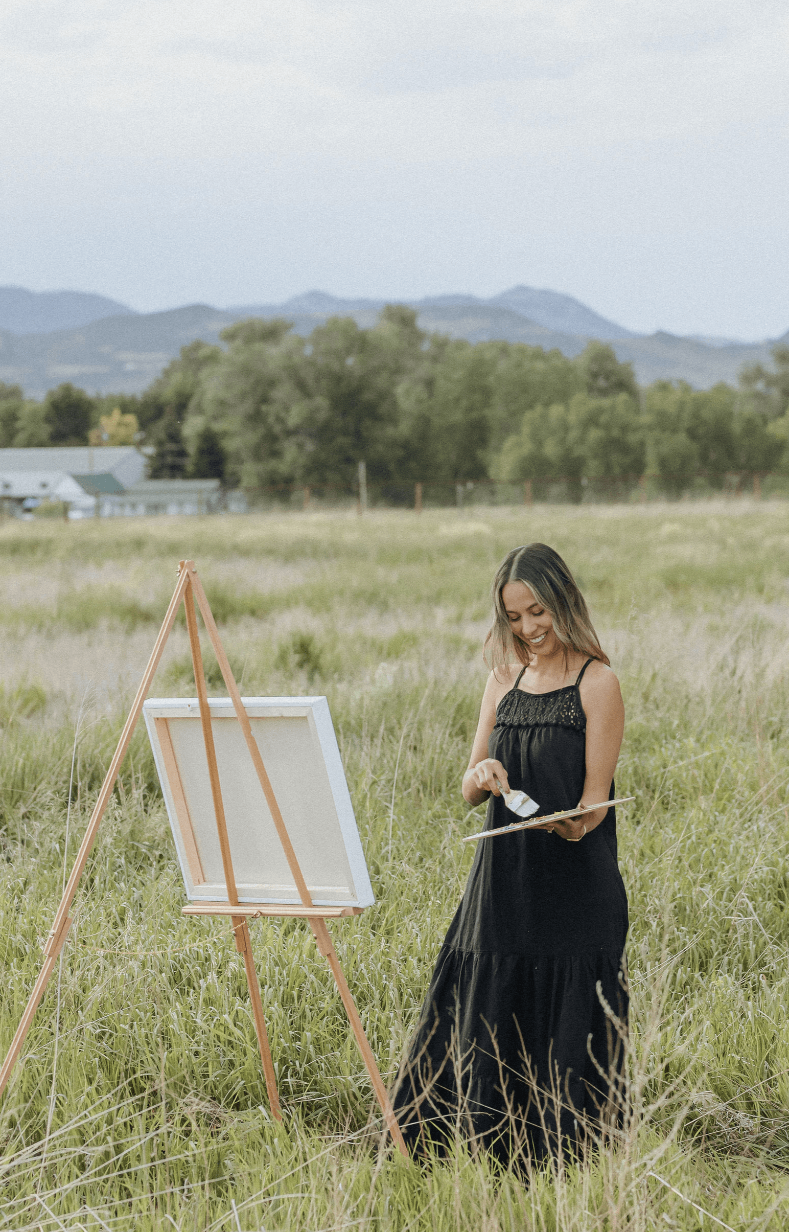Woman in a black dress standing in a grassy field, holding a paintbrush, with an easel nearby, landscape background.