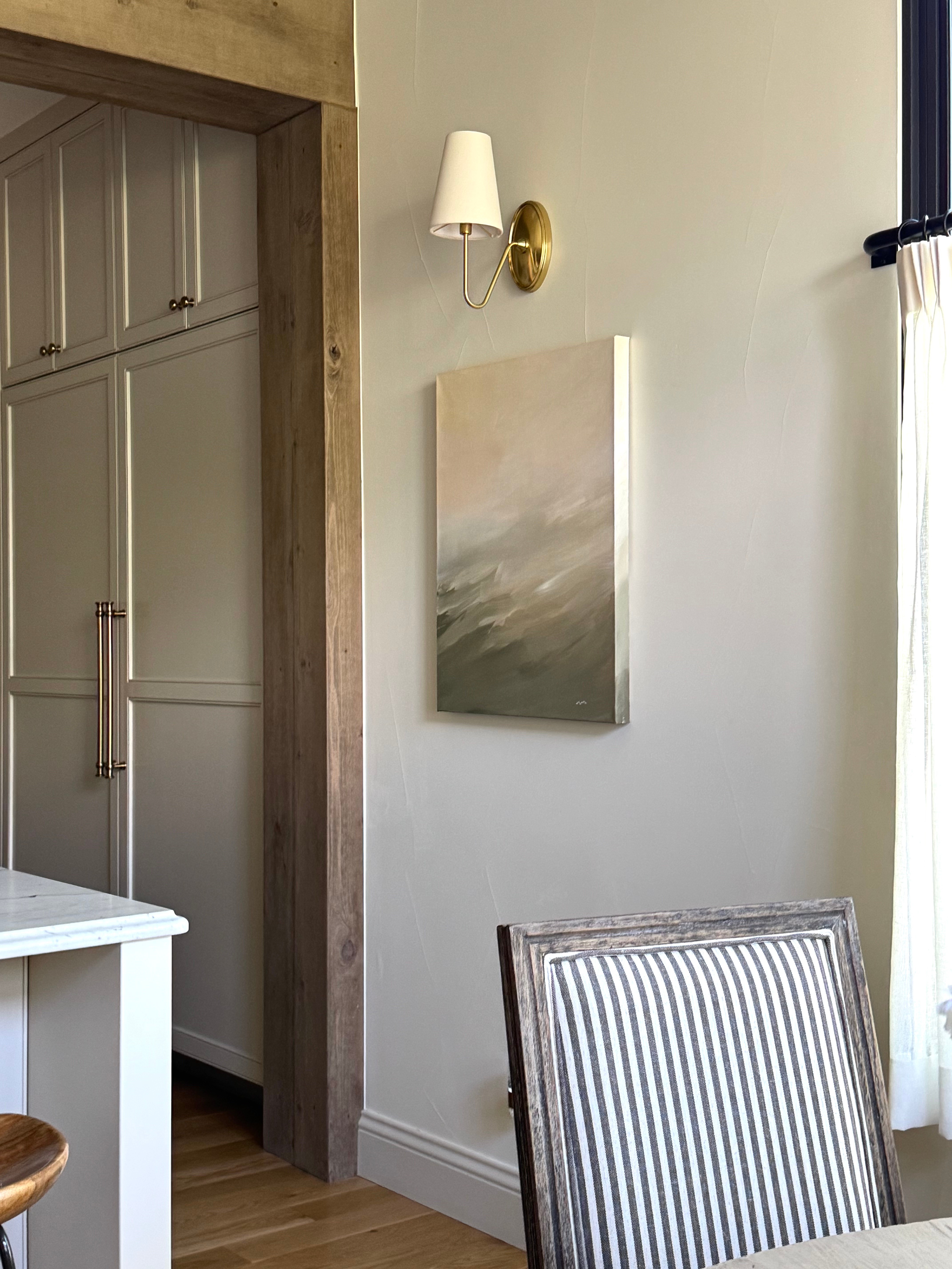 Wall with a white abstract painting, a wall-mounted lamp, and a window with curtains, part of a dining area.
