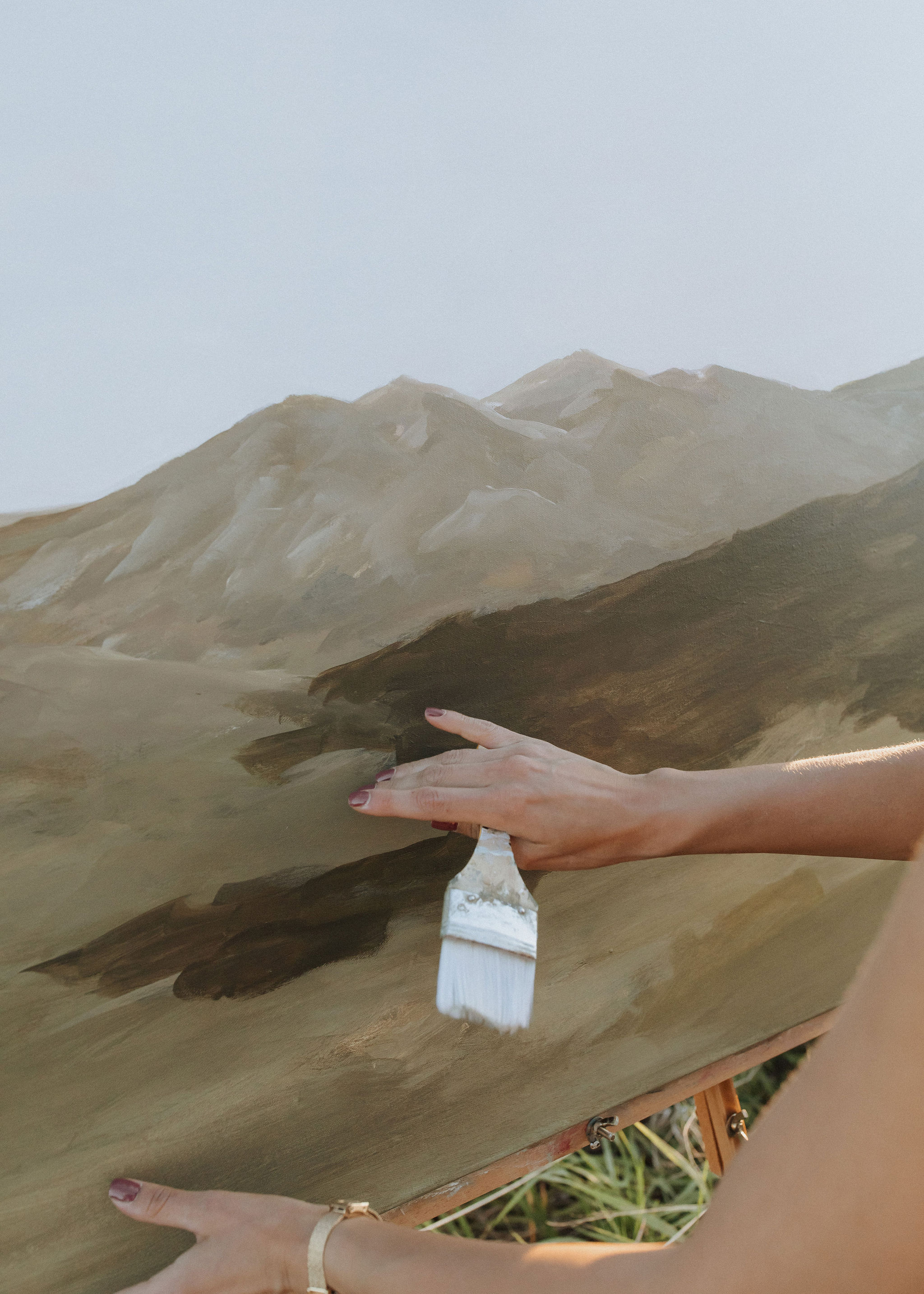 Person's hand holding a paintbrush painting a mountain landscape on canvas, with grass visible at the bottom.