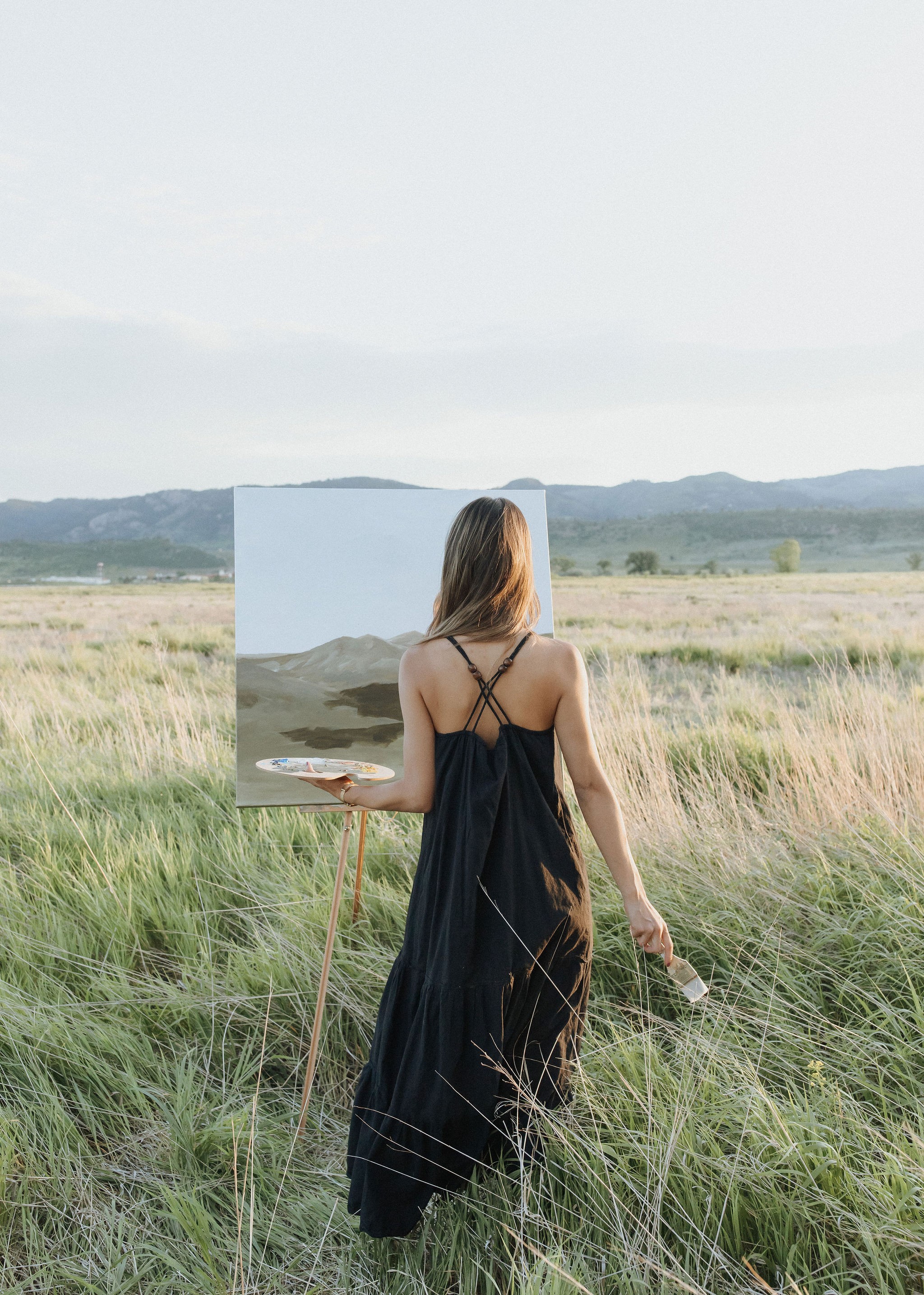 Person in a black dress painting outdoors on a canvas with a landscape background, grassy field, and distant hills.
